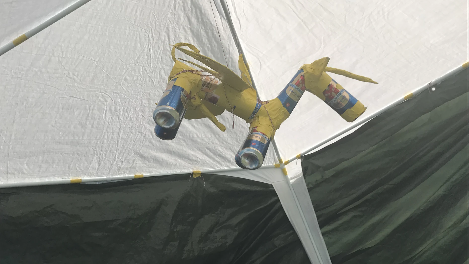 A sculpture resembling a dragon hangs from the ceiling of a tent. It is constructed from yellow material and aluminum cans, with two cans attached as eyes and others forming parts of its body, all secured with tesa tape. The tent is green and white. (This text has been generated by AI)