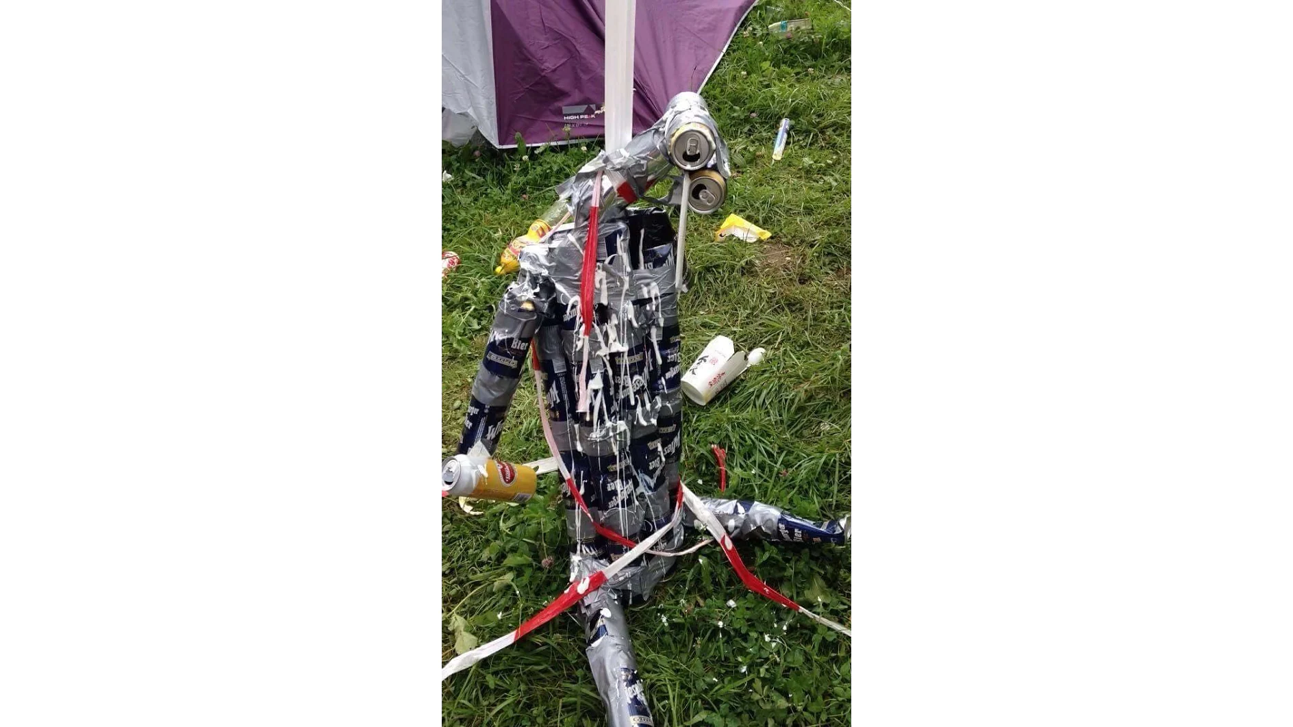 A structure made of stacked beer cans, tesa tape, and string stands on grass. The cans are mostly dark blue and some silver. White spray foam covers parts of the structure. Nearby are more cans and plastic bottles. (This text has been generated by AI)