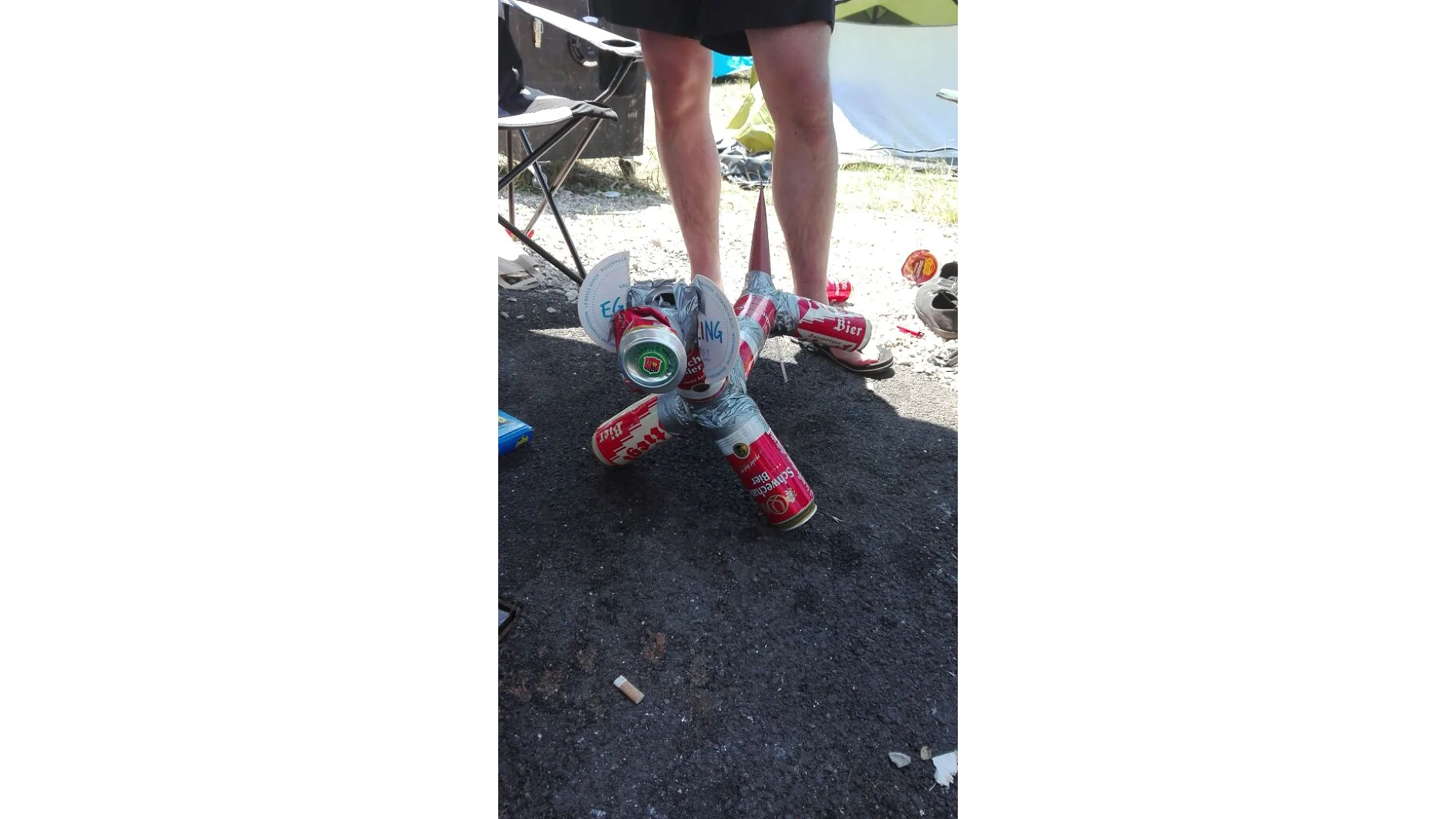 A person stands next to a makeshift creation made of red and silver beer cans held together with tesa tape. The structure is on a paved surface, and various camping items appear in the background. Only the persons legs are visible. (This text has been generated by AI)
