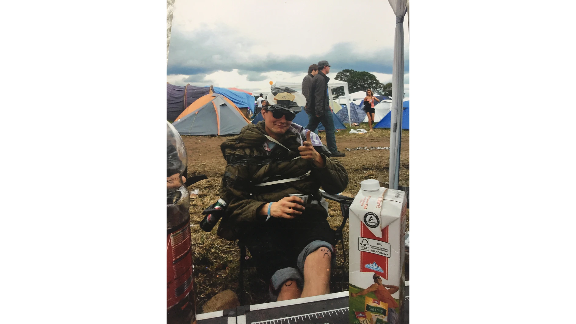 Person sitting on a folding chair at an outdoor event. They are wearing sunglasses and a hat, with multiple layers of clothing. Holding a drink and pointing. Tents are pitched on a grassy field in the background under a cloudy sky. (This text has been generated by AI)