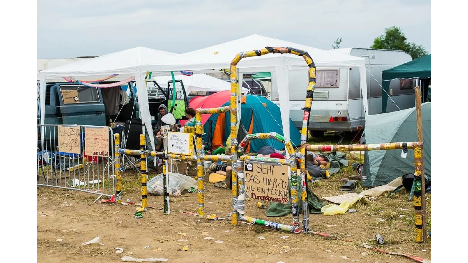 A campsite with tents and a van is surrounded by a makeshift barrier. The ground is covered with trash and camping gear. A sign is secured to the barrier using tesa tape. In the background, there are white canopies and a trailer under a cloudy sky. (This text has been generated by AI)