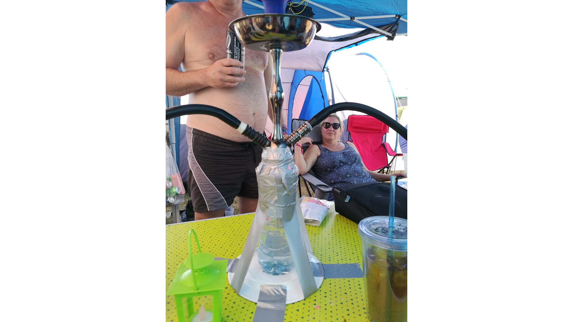 A homemade hookah sits on a yellow table, with tesa tape securing a plastic bottle. Two people are in the background, one seated and wearing sunglasses. An open tent is visible, along with a green lantern and a drink on the table. (This text has been generated by AI)