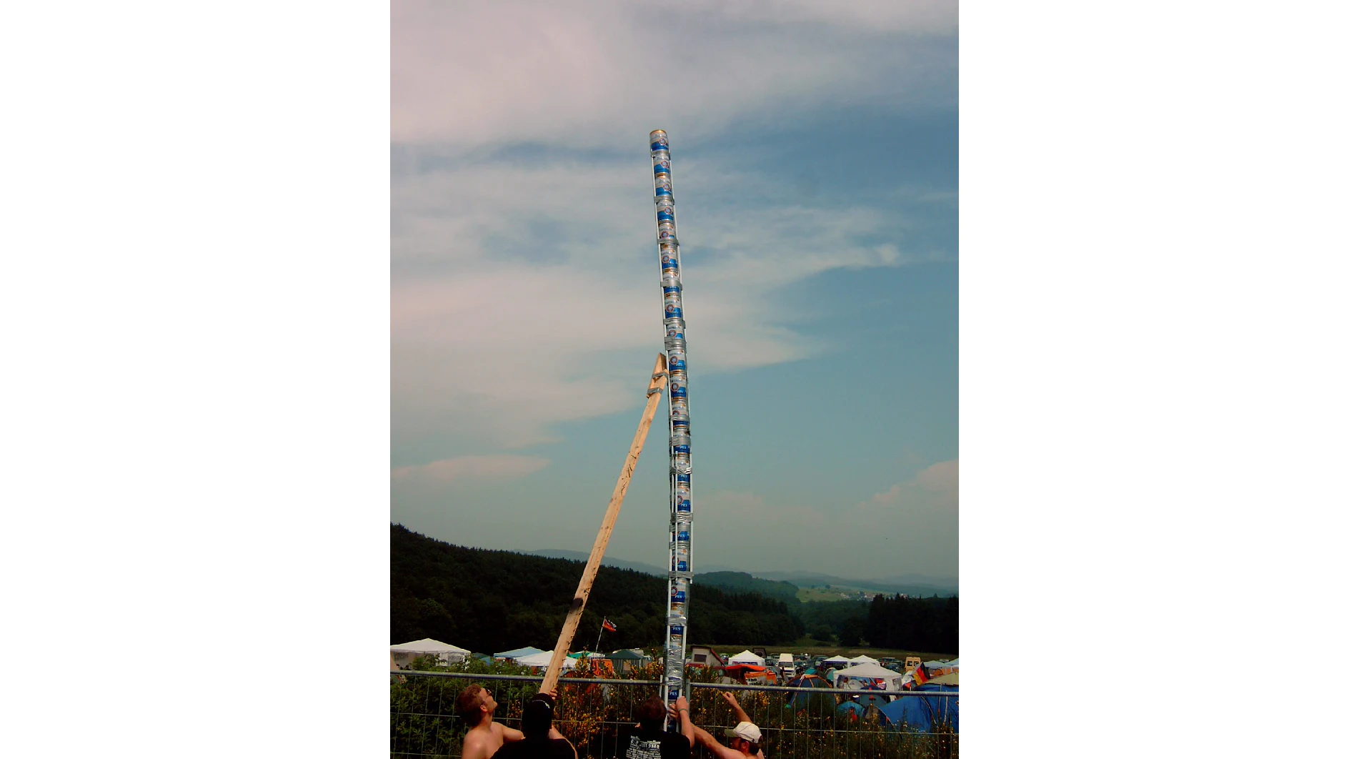 Two people are balancing a tall stack of beer cans with wooden and metal poles using tesa tape. The background features a campground with tents and distant hills under a partly cloudy sky. (This text has been generated by AI)