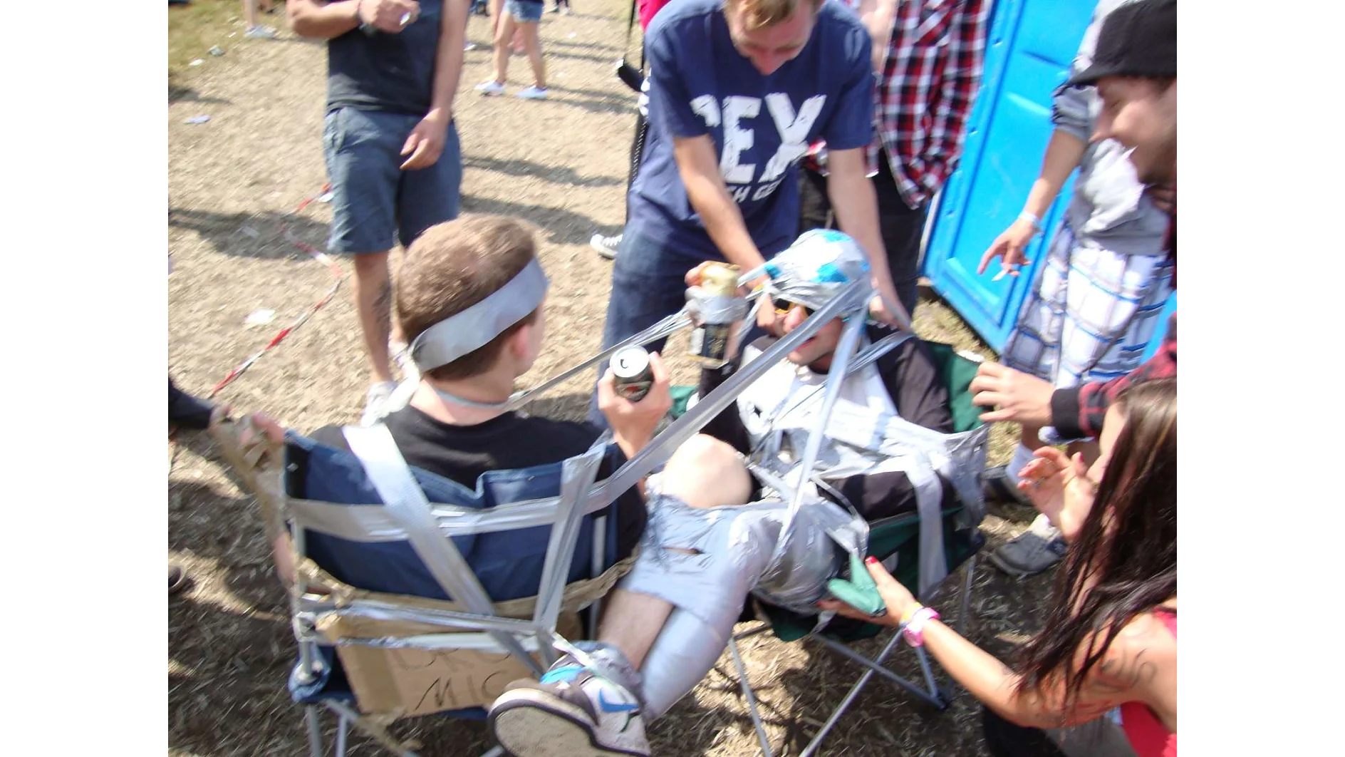 A group of people appears to be using tesa tape to secure two men to chairs at an outdoor event. One man holds a drink can while another person points at him. A portable toilet is visible in the background. The scene seems playful and informal. (This text has been generated by AI)