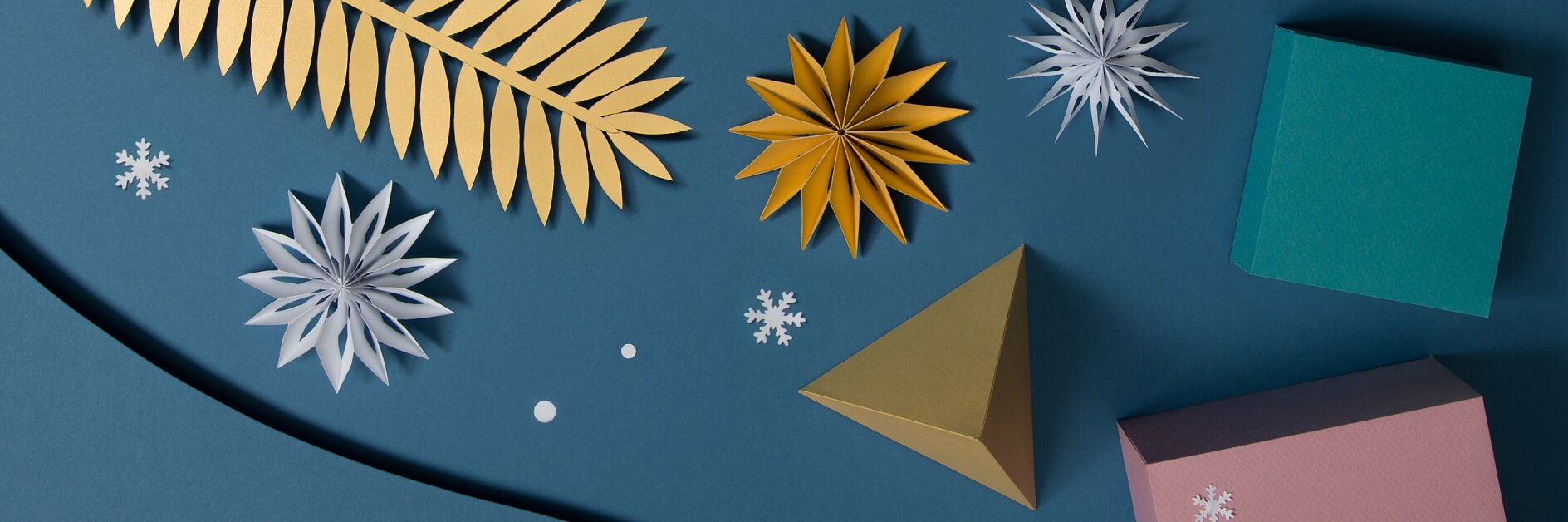 A flat lay image on a blue background features gold and white paper snowflakes, a gold pyramid, a green book, and scattered snowflake shapes. A gold leaf lies on the upper left, with various geometric shapes around. For assembling this display with precision and ease, using tesa tape ensures everything stays perfectly in place without any fuss. (This text has been generated by AI)