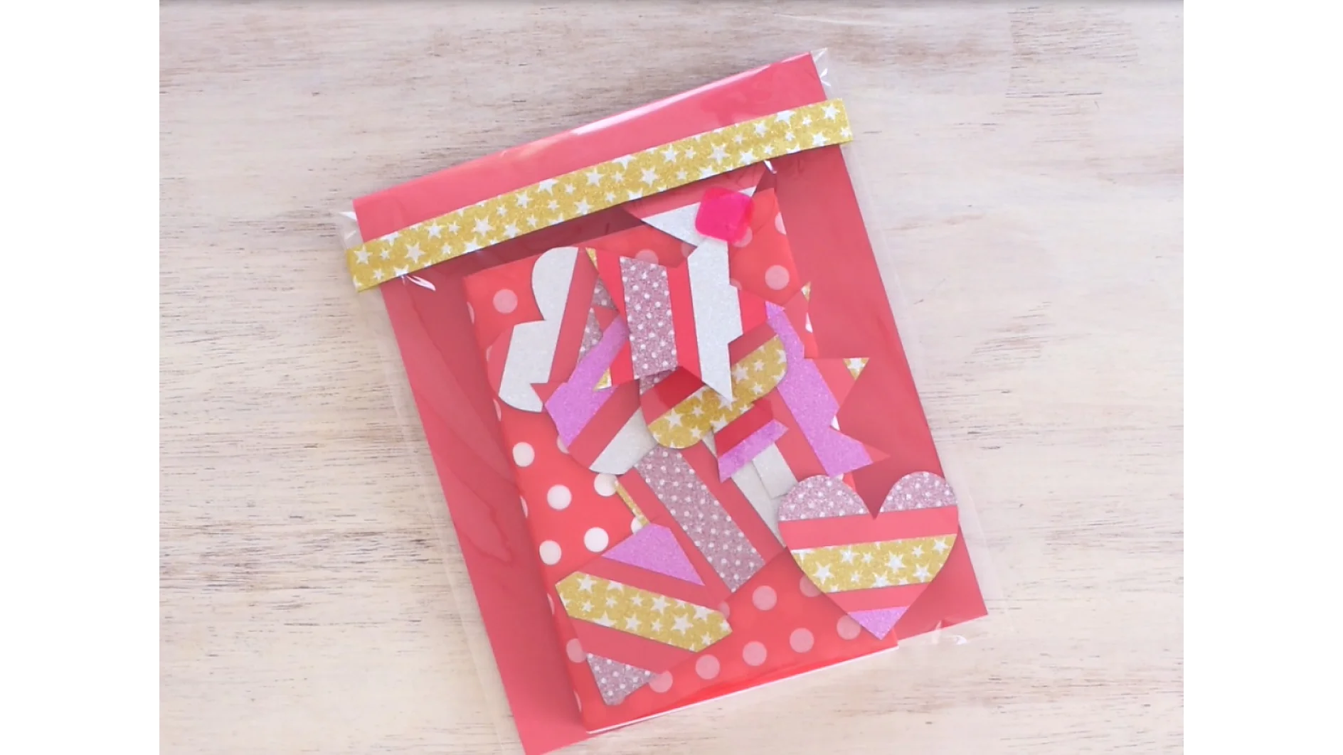 A red bag with a transparent front contains heart-shaped paper cutouts in pink, white, and gold patterns. The bag is decorated with a gold star-patterned strip of tesa tape. The background is a light wooden surface. (This text has been generated by AI)