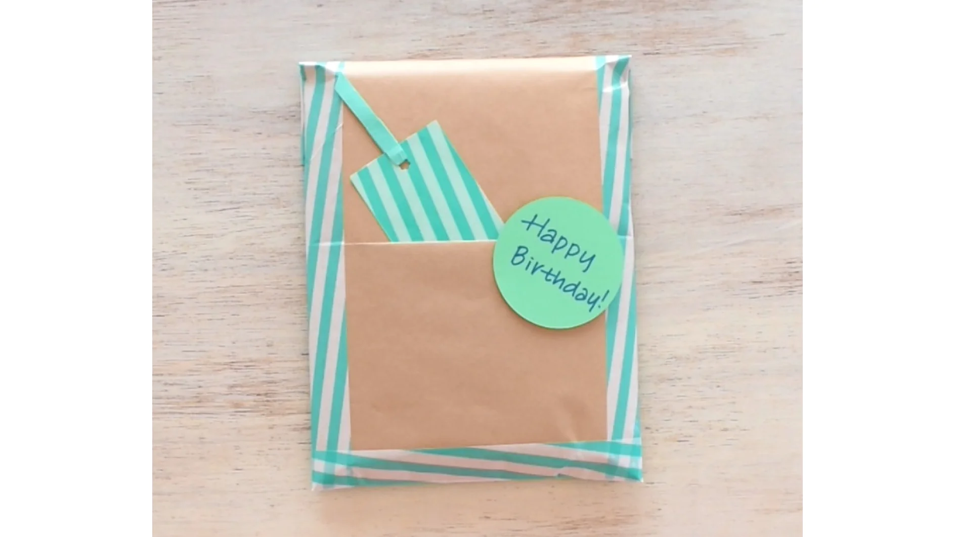 A rectangular gift is wrapped in brown paper with light blue and white striped tesa tape along the edges. A matching blue striped tag is tucked in. A round green tag with Happy Birthday! written on it is attached to the package. (This text has been generated by AI)