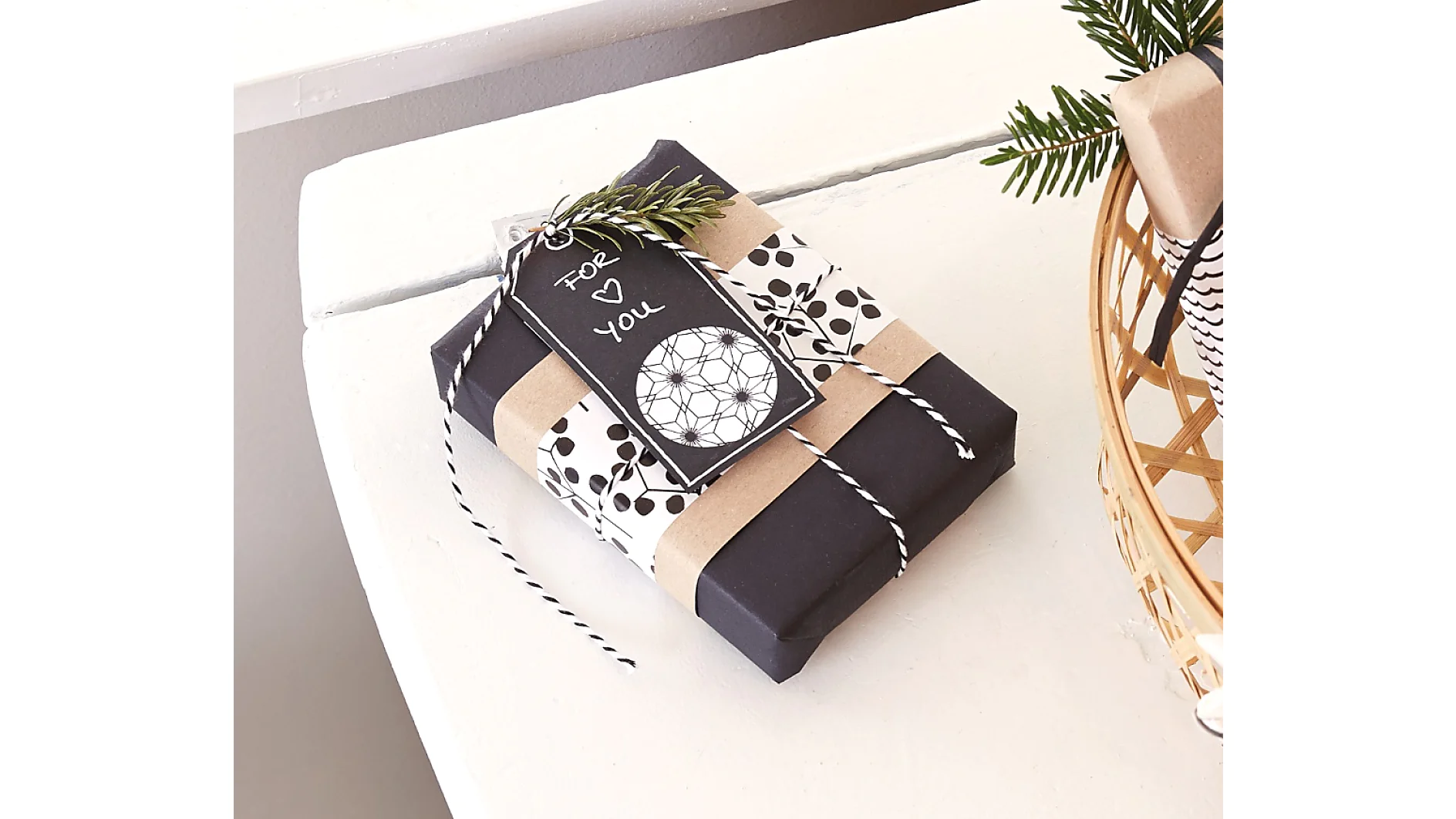 Gift wrapped in black paper with white and brown decorative bands, secured with tesa tape, and tied with black and white twine. A tag featuring a heart and the words For You is attached using tesa tape. A small branch of greenery is tucked under the twine. (This text has been generated by AI)