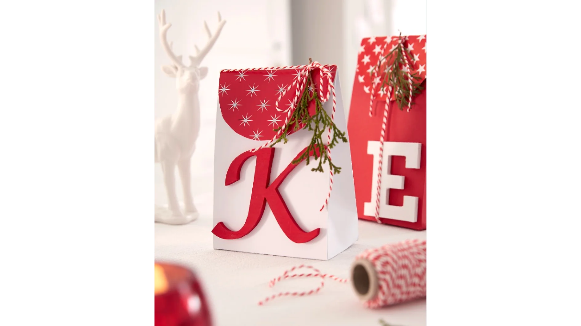 Decorative gift bags with large red letters K and E on a white table. One bag is accented with a star pattern and greenery. Red and white twine is nearby, secured by tesa tape. A white reindeer figurine is in the background. (This text has been generated by AI)
