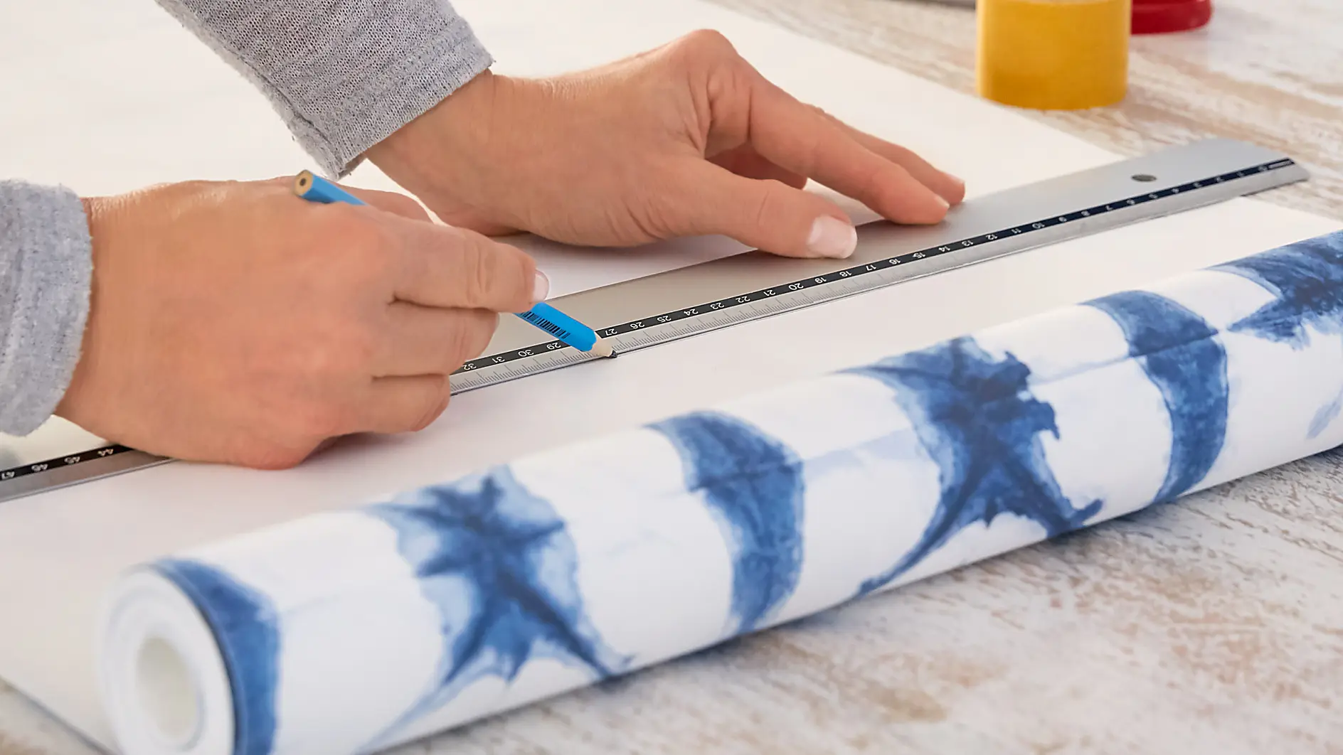 Hands measuring and marking wallpaper with a ruler and pencil on a wooden table. A roll of blue and white patterned wallpaper, scissors, and tesa tape are visible.