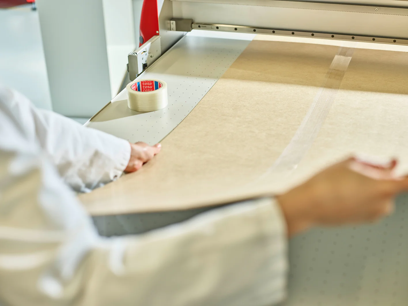 Person in a white coat placing large sheets of brown paper on a cutting machine. A roll of clear tesa tape is on the table beside them. The setting appears to be a professional workspace or workshop. (This text has been generated by AI)