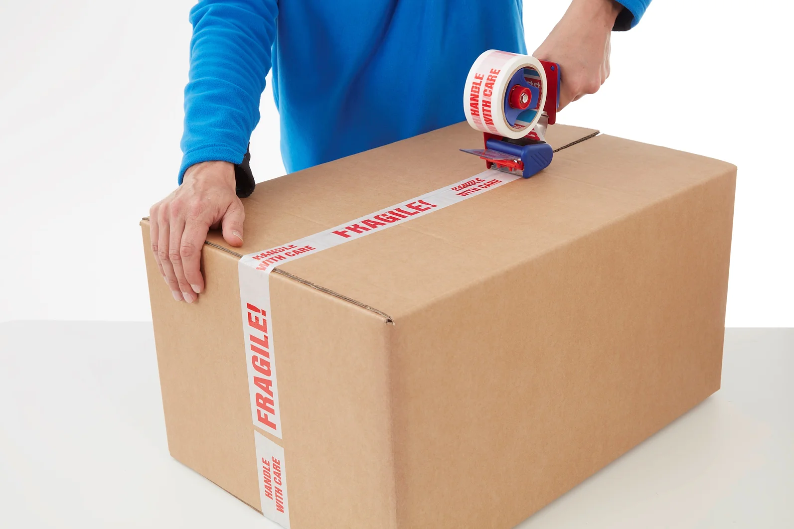 6120 printed tape A person in a blue shirt is sealing a large cardboard box with white tesa tape labeled FRAGILE! in red letters. The box is on a white surface, and the tesa tape dispenser is blue with a red handle. (This text has been generated by AI)