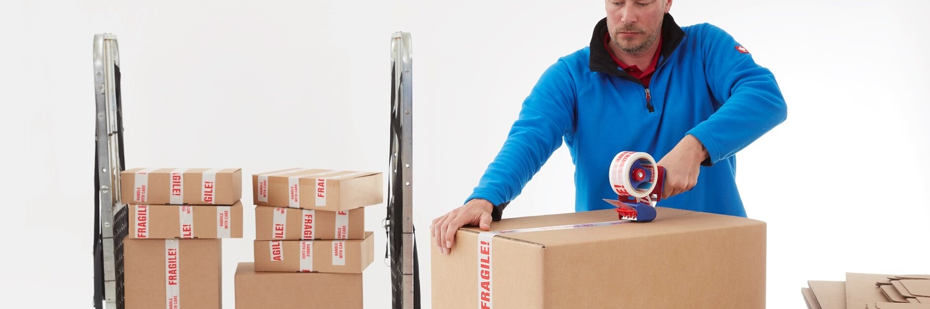 A man in a blue sweater uses tesa tape on a cardboard box labeled Fragile on a white table. Behind him is a cart stacked with similar boxes. A few flattened boxes are on the table. The background is plain white. (This text has been generated by AI)
