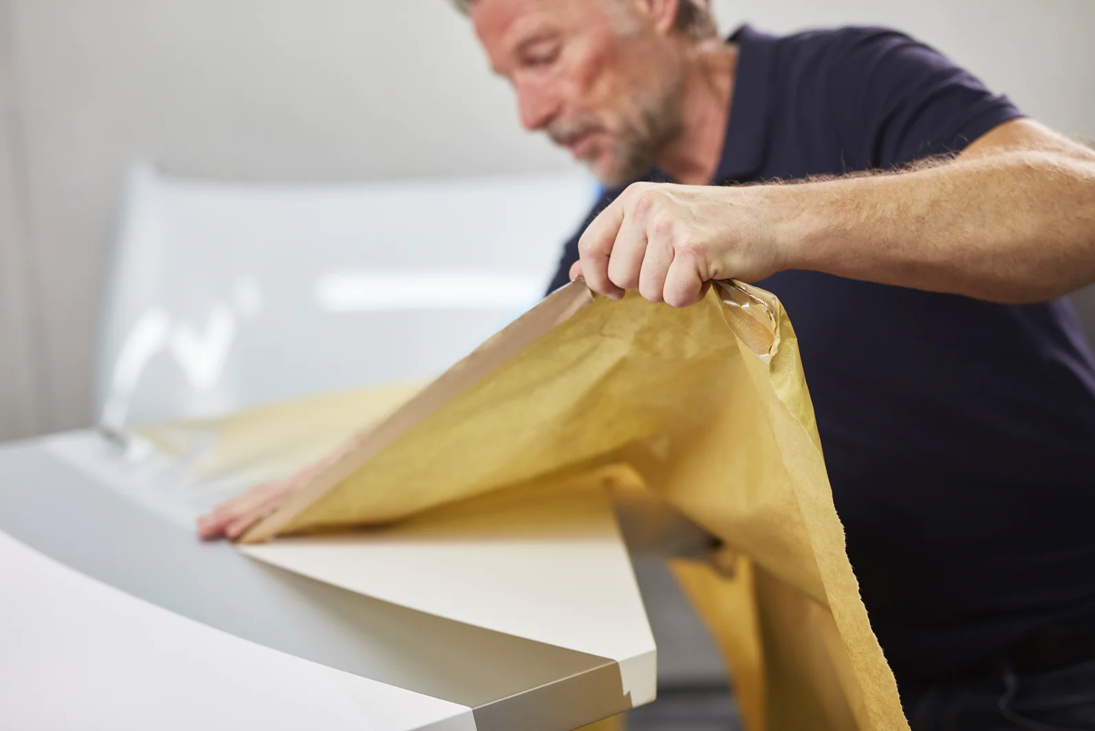 A man in a black shirt carefully peels back brown paper covering a white surface, possibly preparing or inspecting a product. The background is blurred. (This text has been generated by AI)