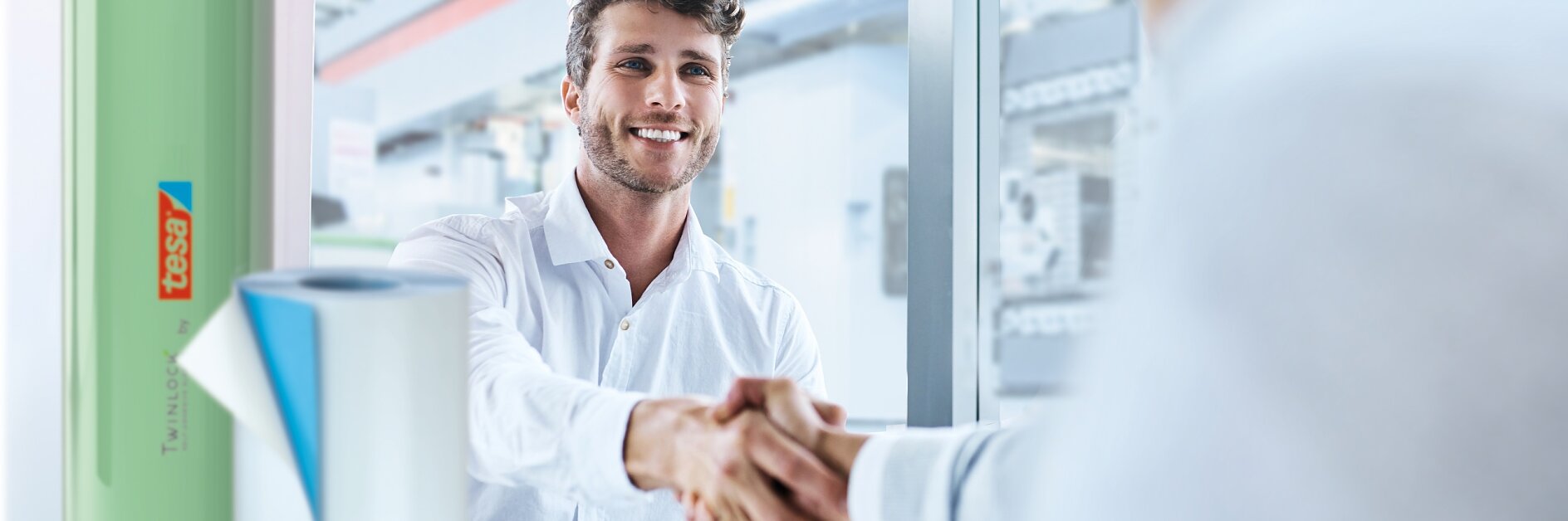 Two people in a bright office setting are shaking hands across a desk. One person is smiling, wearing a white shirt. On the desk, there is a green product cylinder with tesa tape branding next to it. A window shows an industrial interior background. (This text has been generated by AI)