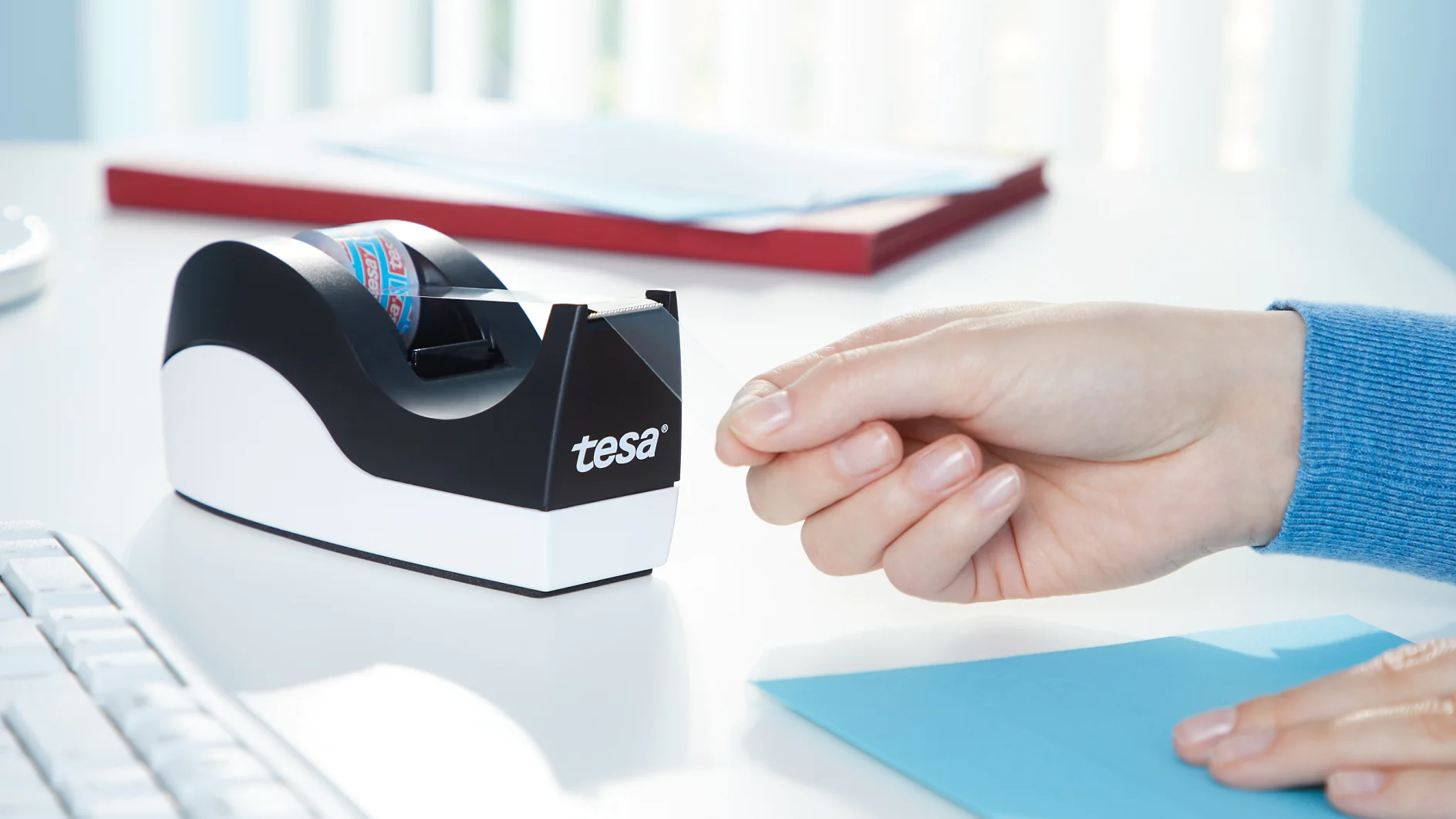 A hand is reaching for a piece of tesa tape from a black and white tape dispenser labeled tesa on a white desk. A keyboard, blue paper, and a red notebook are visible in the background. (This text has been generated by AI)