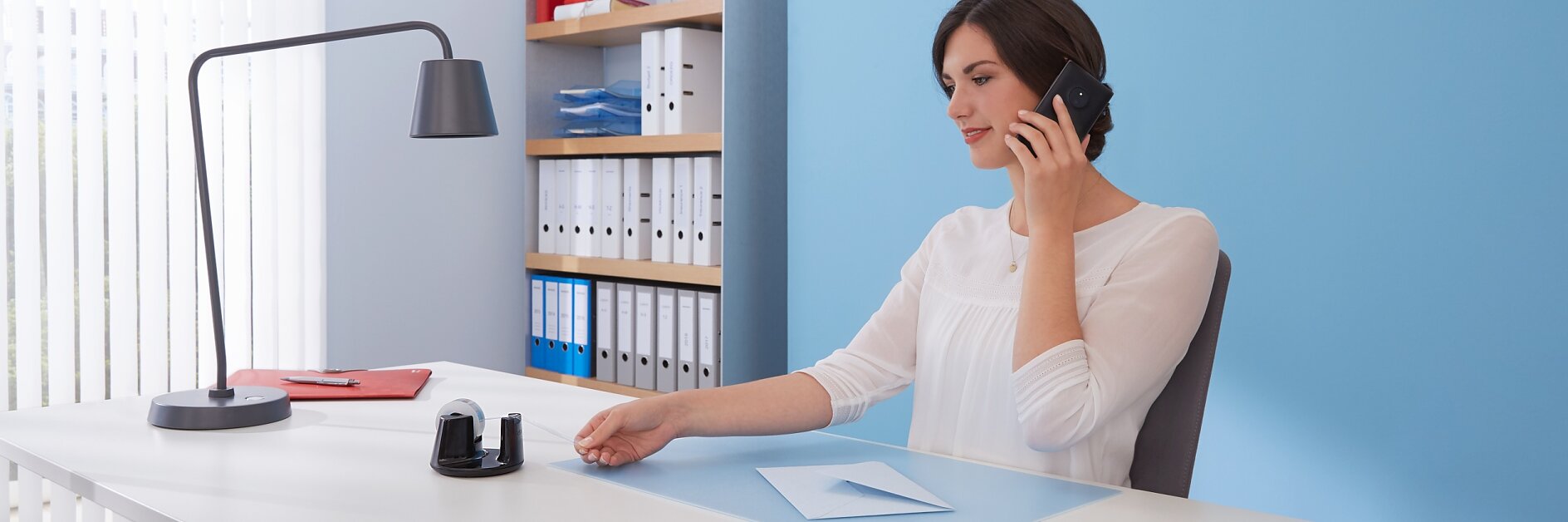 A woman is sitting at a desk in an office, talking on a smartphone. She wears a white blouse and reaches toward a tesa tape dispenser. The desk has a lamp, document, and a red folder. Behind her is a shelving unit with binders and folders. (This text has been generated by AI)