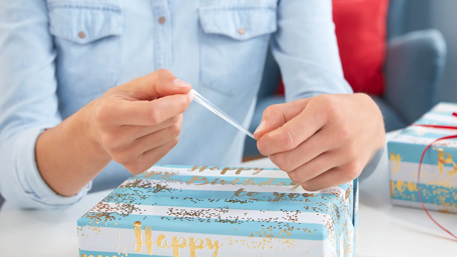 A person wearing a light blue shirt is using clear tesa tape to wrap a gift. The gift is covered in blue and white wrapping paper with gold text that reads Happy Birthday to you. Another wrapped gift is partially visible in the background. (This text has been generated by AI)