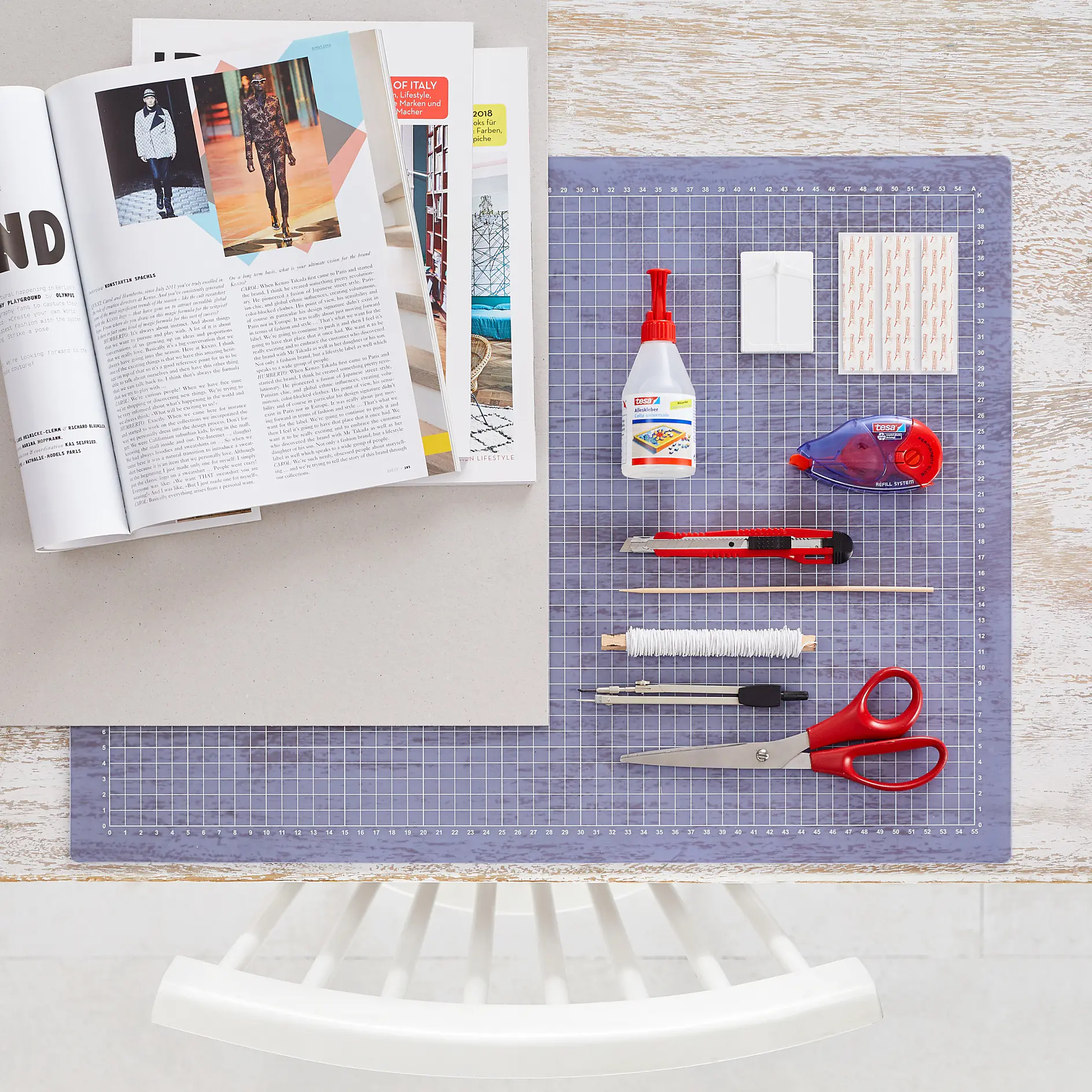 A top-down view of a crafting workspace. On the left, an open magazine. On the right, a grid mat with supplies: glue, tesa tape adhesive strips, correction tesa tape, pens, a pencil, sewing needle, thread spool, scissors, utility knife, and a cutting tool. A chair is visible below. (This text has been generated by AI)