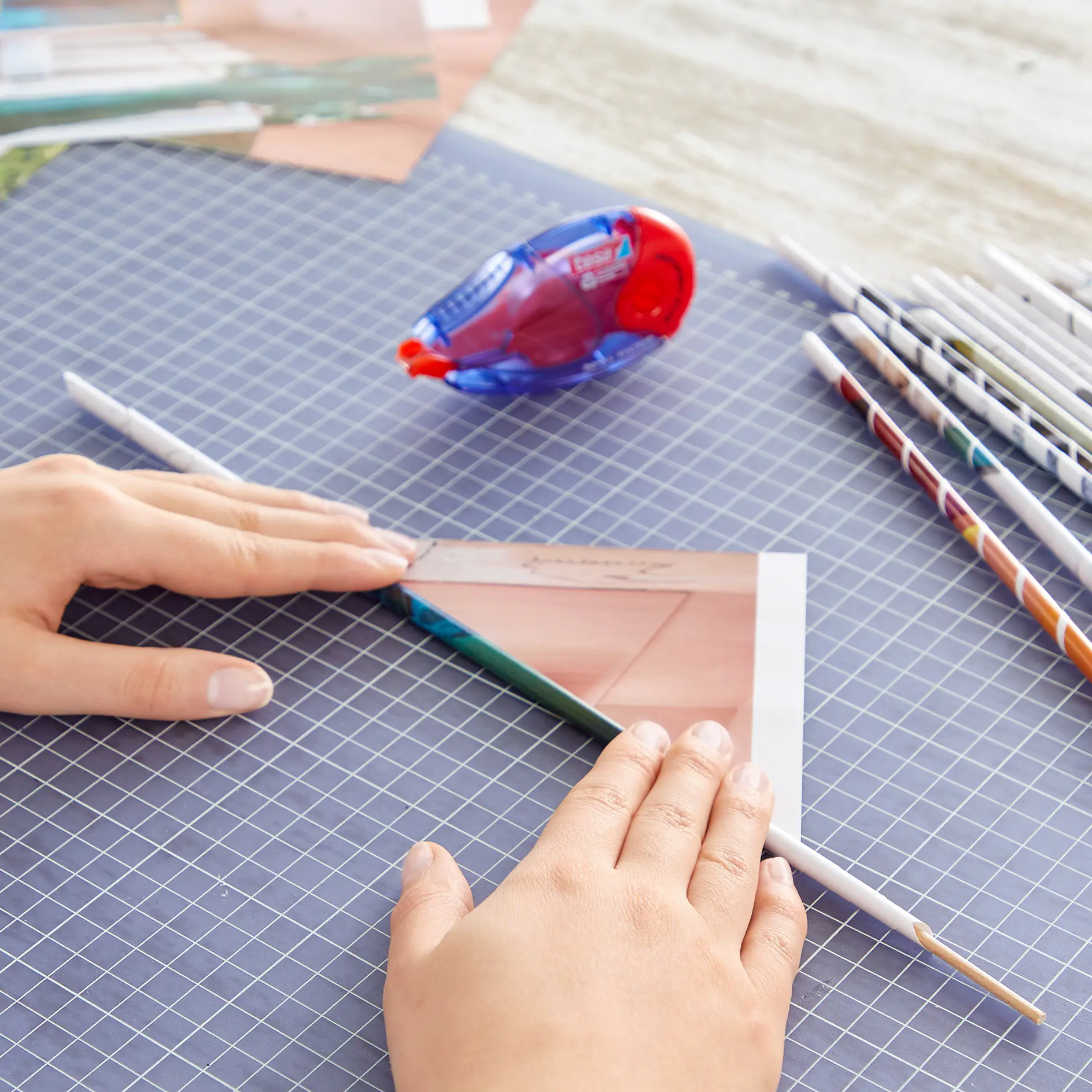 Hands rolling a sheet of paper into a tight tube on a grid-patterned surface. A blue and red adhesive dispenser is nearby, along with several completed paper tubes on the right. Several rolls of tesa tape are present for securing the tubes tightly. (This text has been generated by AI)