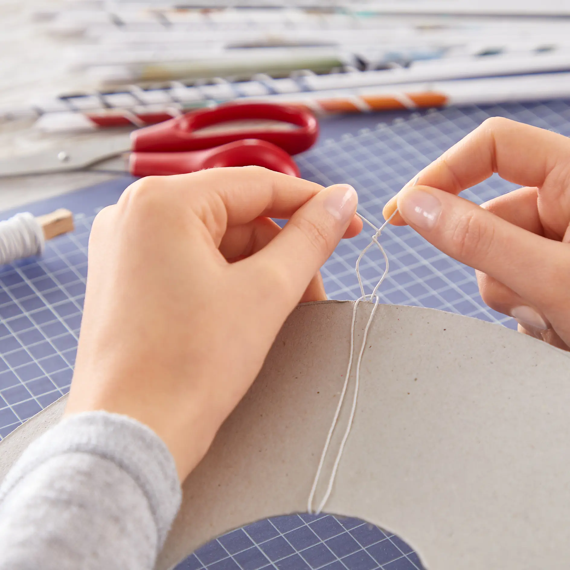 Close-up of hands tying string around a piece of cardboard. Scissors and paper rolls are in the background. The surface is covered with a blue cutting mat featuring a white grid pattern, with tesa tape visible nearby. (This text has been generated by AI)