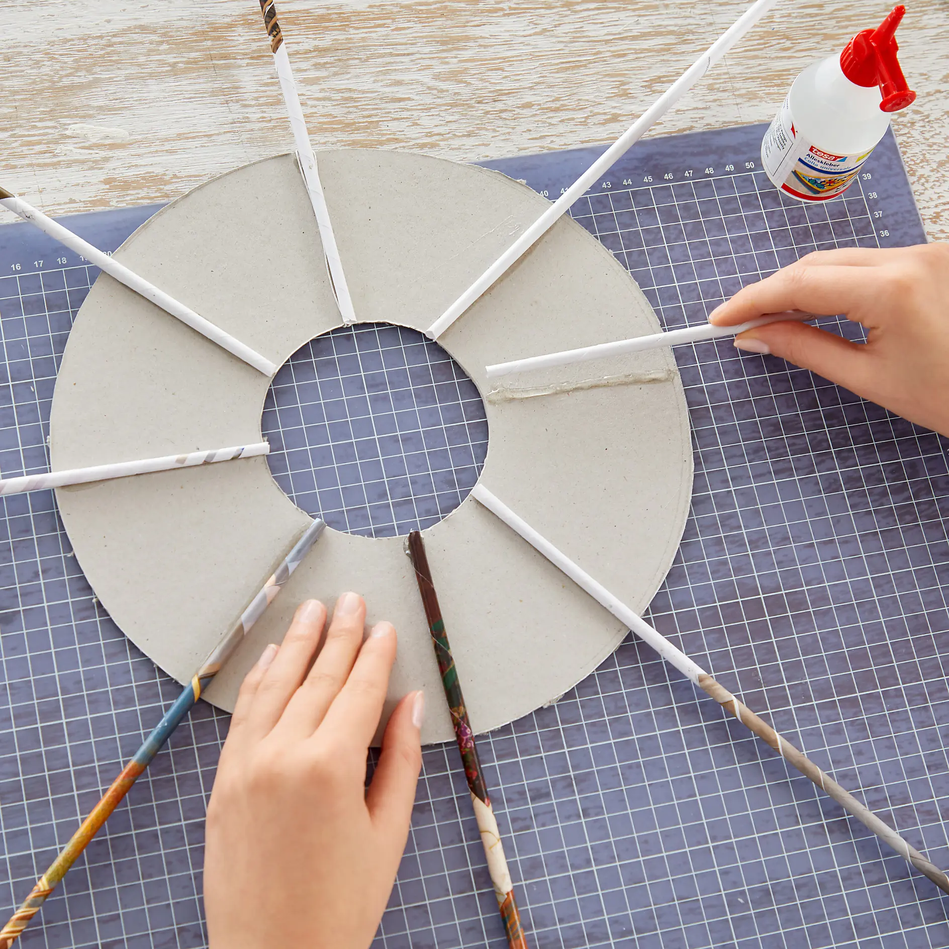 Hands are creating a circular craft with a cardboard base and rolled paper tubes, secured with tesa tape. A small bottle of glue is on the right. A cutting mat with a grid pattern is underneath the project. (This text has been generated by AI)