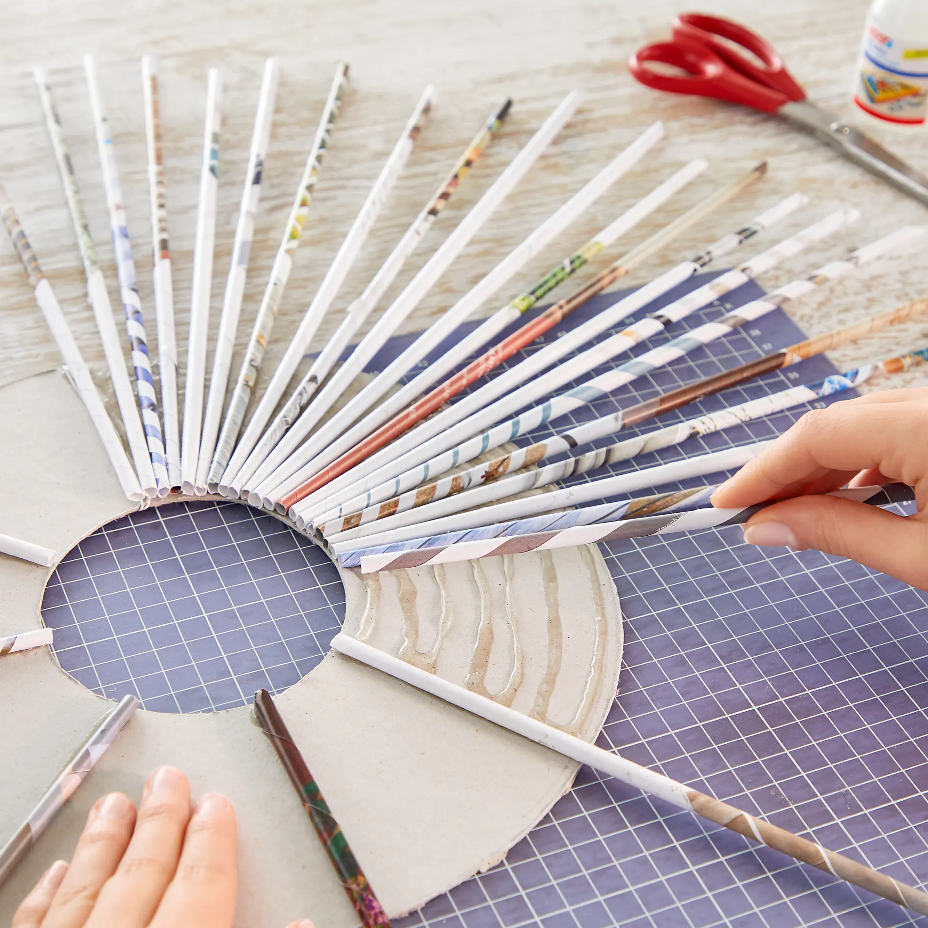 Hands are weaving rolled paper strips onto a circular piece of cardboard. Scissors, glue, and additional rolled strips are in the background on a grid-patterned surface. (This text has been generated by AI)
