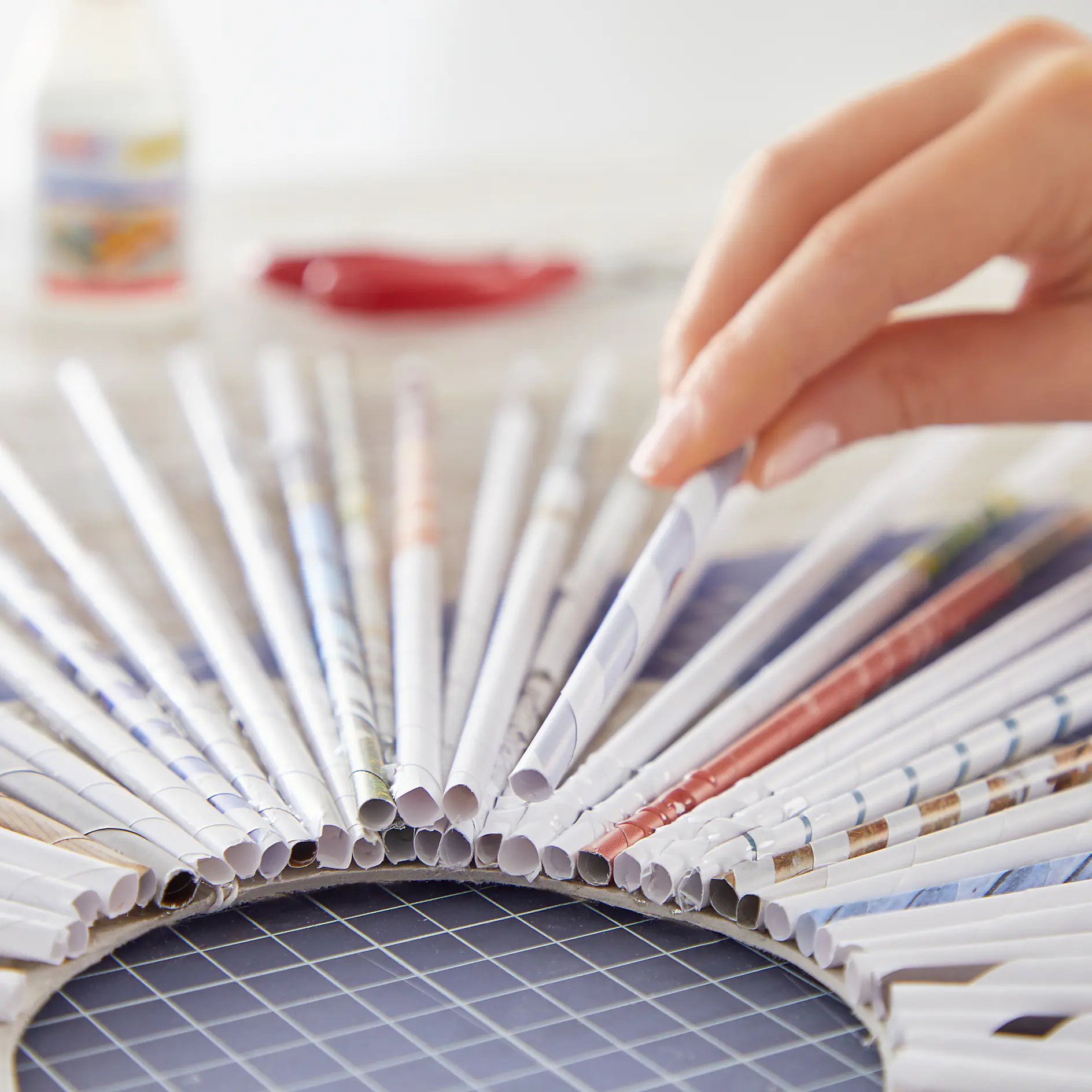 A hand arranging rolled-up strips of colorful paper into a circular pattern on a gridded surface, with tesa tape and crafting materials in the background. (This text has been generated by AI)