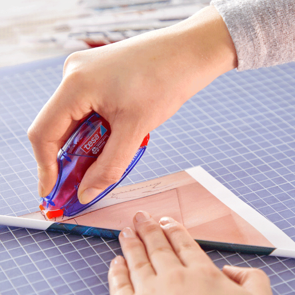A person using a correction tesa tape dispenser to apply adhesive to the corner of a piece of paper placed on a blue cutting mat. The persons hand is pressing the paper flat while guiding the dispenser along the edge. (This text has been generated by AI)