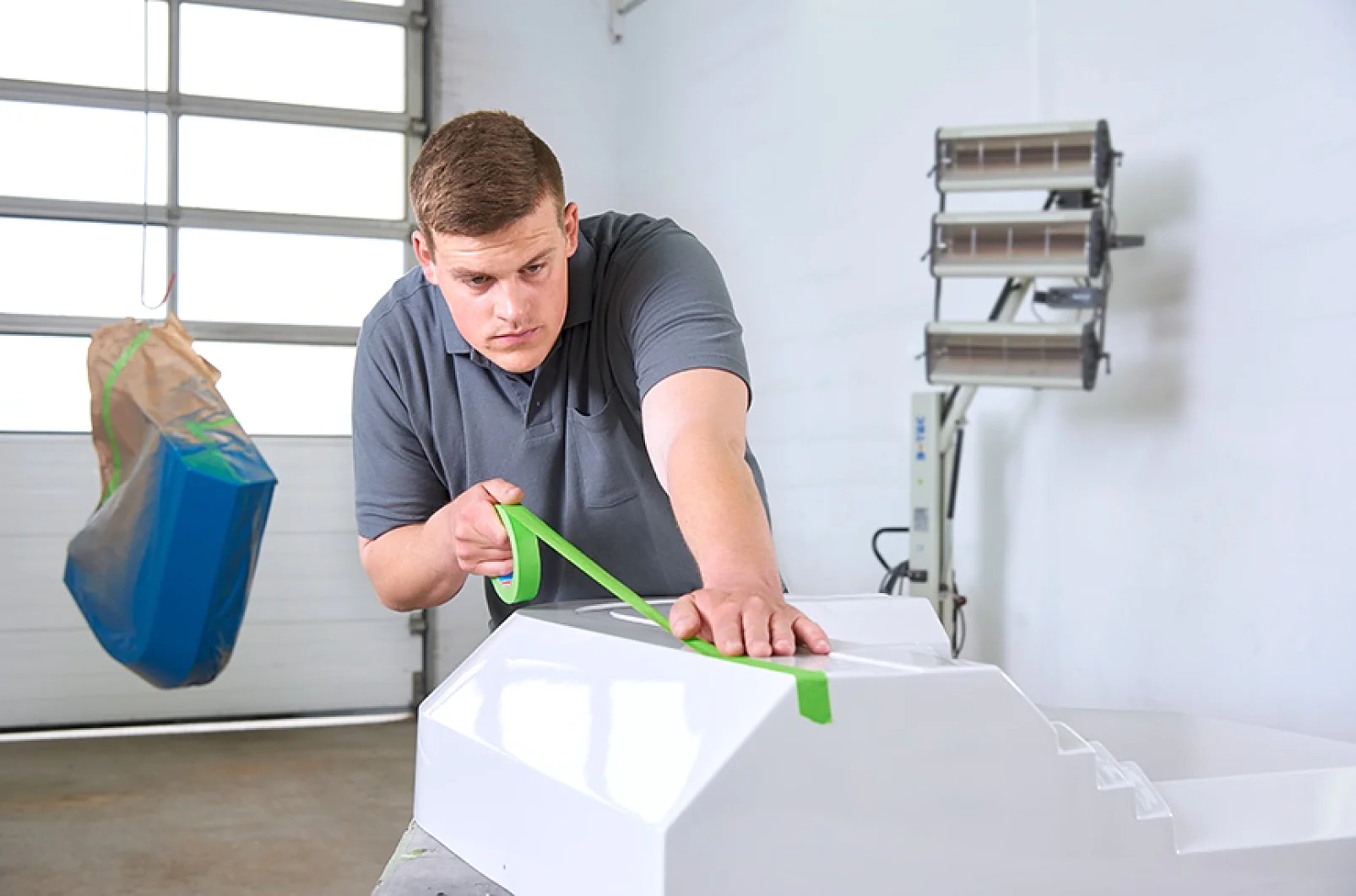 A man in a gray shirt applies green tesa tape to a white object in a garage-like setting. A blue bag hangs on the left, and a set of lamps is on the right. The scene suggests preparation for painting or similar work. (This text has been generated by AI)