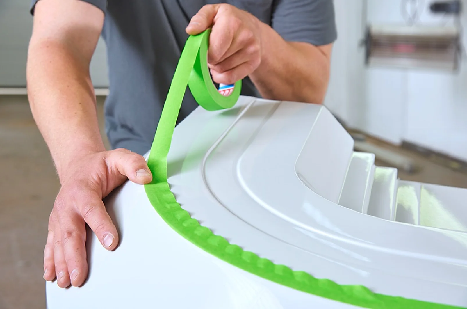 A person applying green tesa tape to a curved white surface, possibly for painting or coating purposes. The scene appears to be indoors, with tools visible in the blurred background. (This text has been generated by AI)