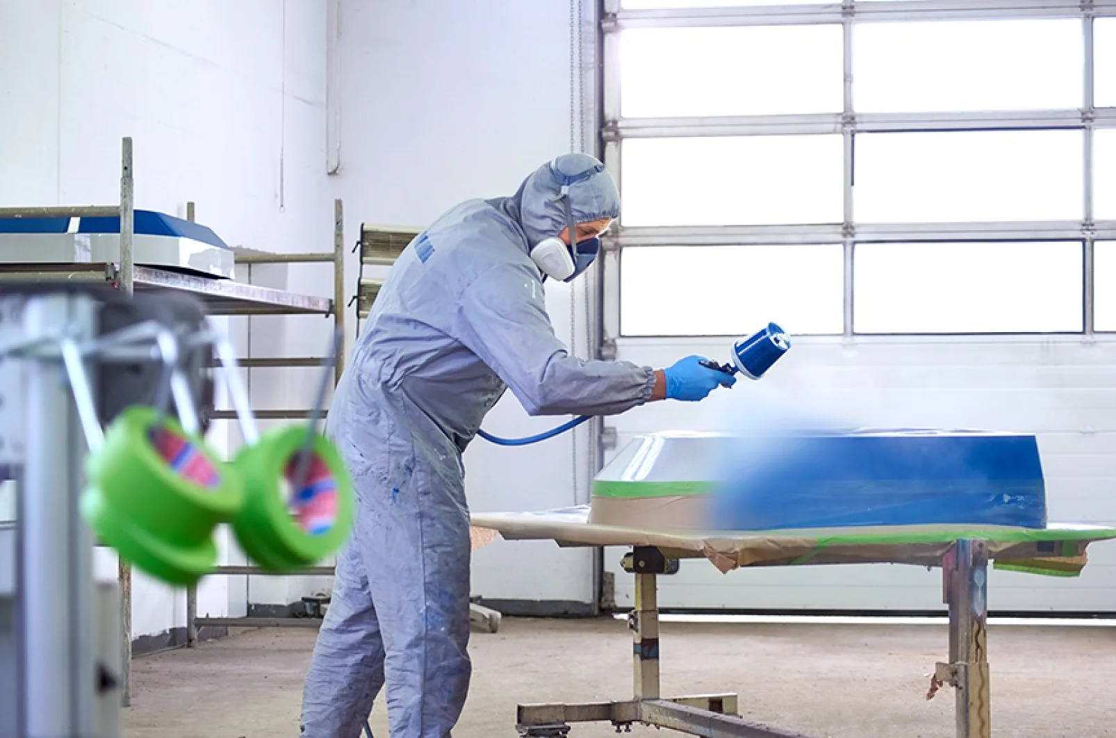 A person wearing protective gear, including a respirator and goggles, is using a spray gun to paint a large metallic object in a workshop. The area has bright lighting and visible tools and tesa tape in the background. (This text has been generated by AI)