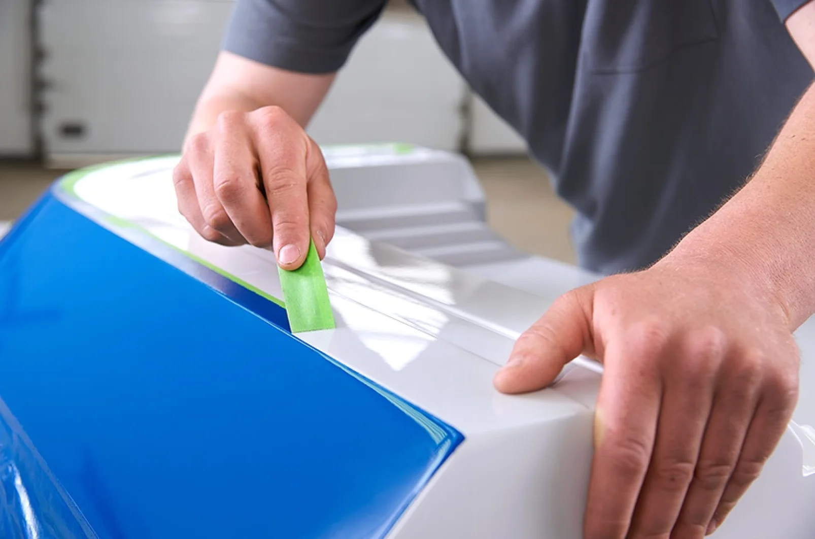 A person applies green tesa tape to the edge of a blue and white surface, possibly preparing it for painting or protection. The persons arms are visible, and they are wearing a gray short-sleeved shirt. (This text has been generated by AI)