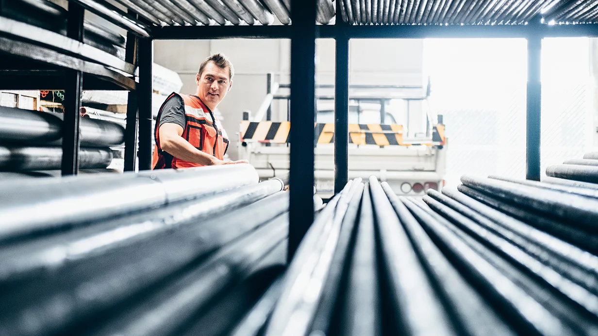 tesa-adhesive-tape-solutions-for-metal-industry A person wearing an orange safety vest stands in a warehouse filled with metal pipes. They are looking to the side, surrounded by the stacked pipes on metal racks, with a truck partially visible in the background. (This text has been generated by AI)