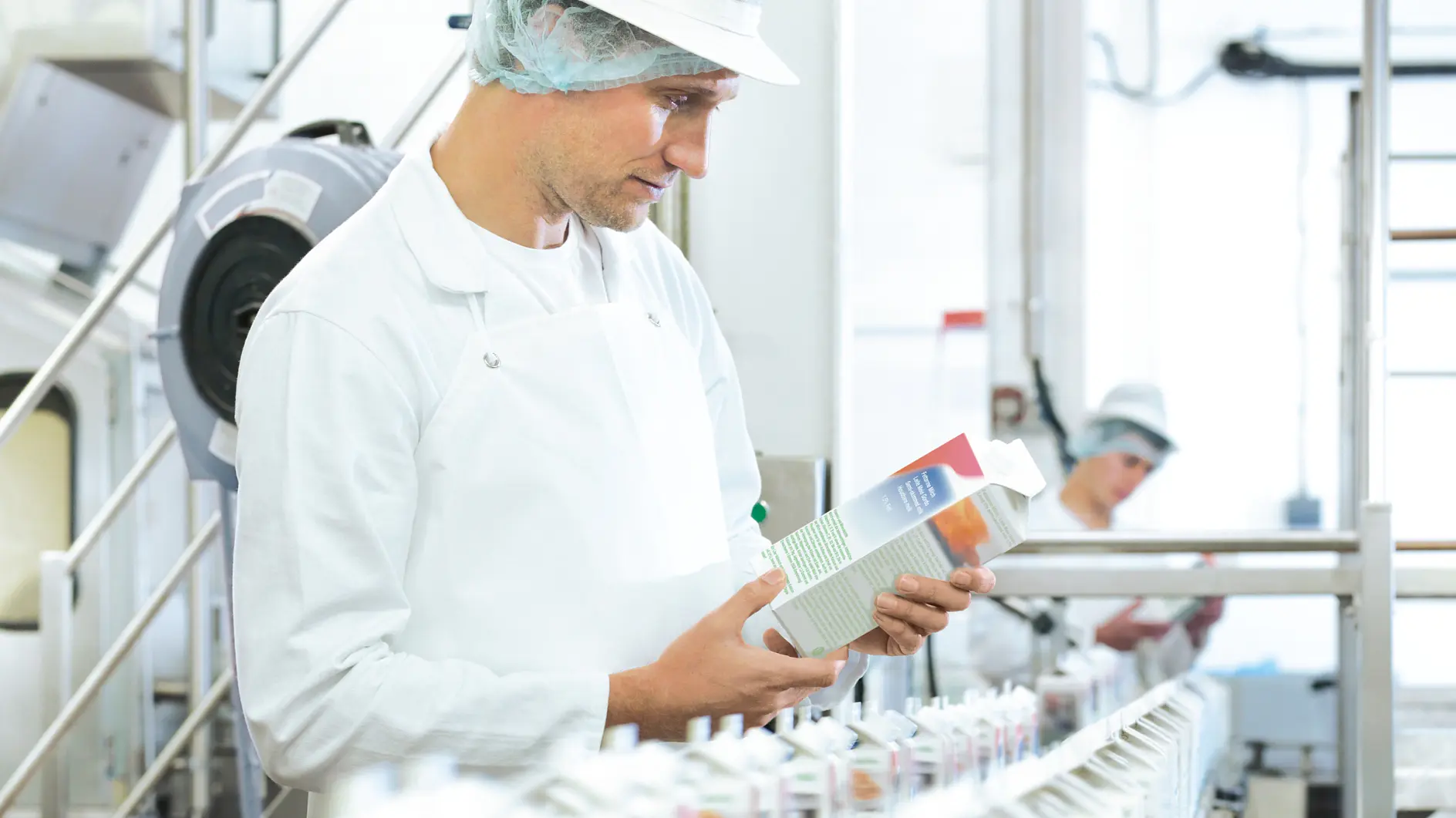 A factory worker in a white apron, hairnet, and cap inspects a carton on an assembly line filled with similar cartons sealed using tesa tape. Another worker in the background is also wearing similar attire in a well-lit, industrial setting. (This text has been generated by AI)