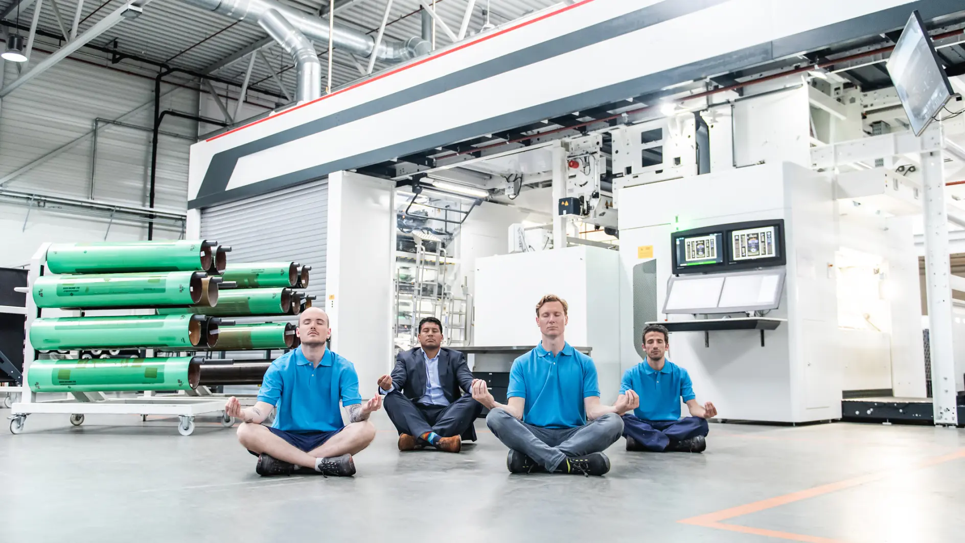 Four people sit cross-legged and meditate on the floor of an industrial workshop. They are wearing blue shirts and are positioned in front of large machinery and rolls of tesa tape material. The setting is well-lit with industrial equipment visible. (This text has been generated by AI)