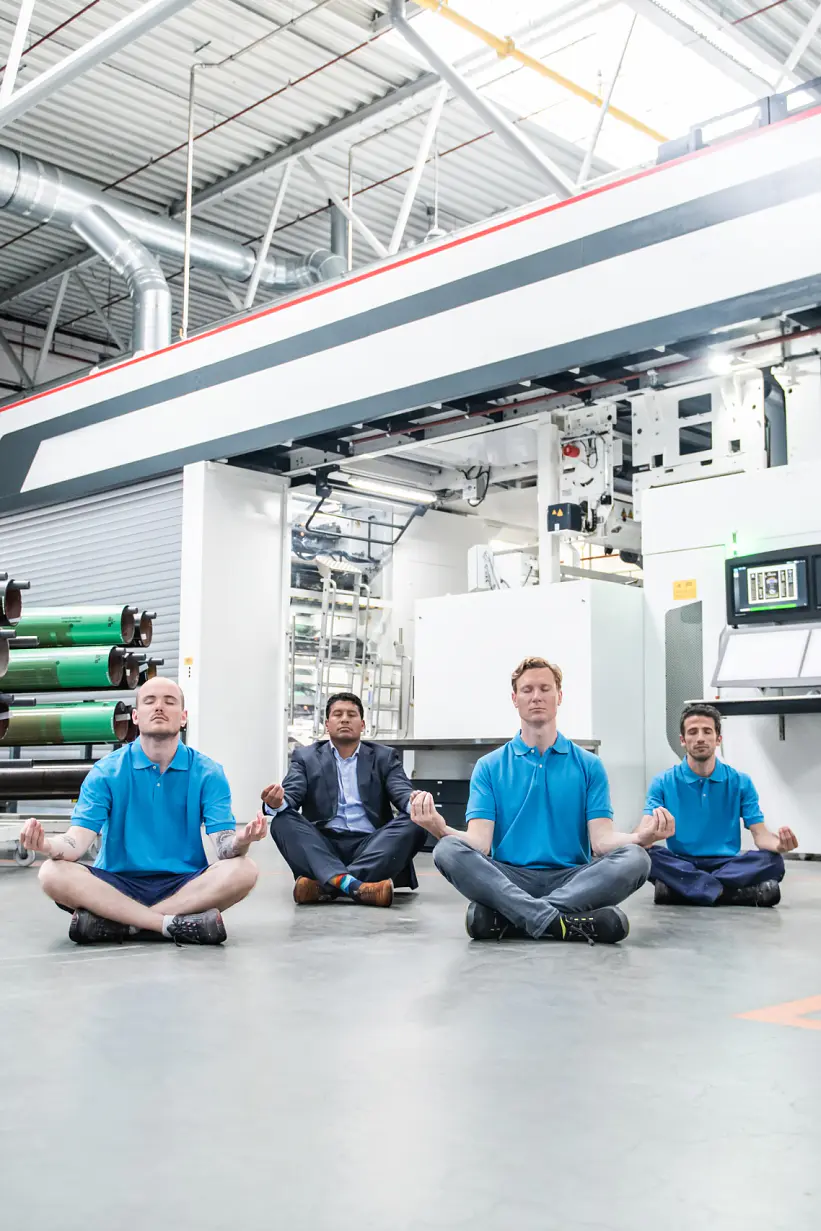 Four people sit cross-legged and meditate on the floor of an industrial workshop. They are wearing blue shirts and are positioned in front of large machinery and rolls of tesa tape material. The setting is well-lit with industrial equipment visible. (This text has been generated by AI)