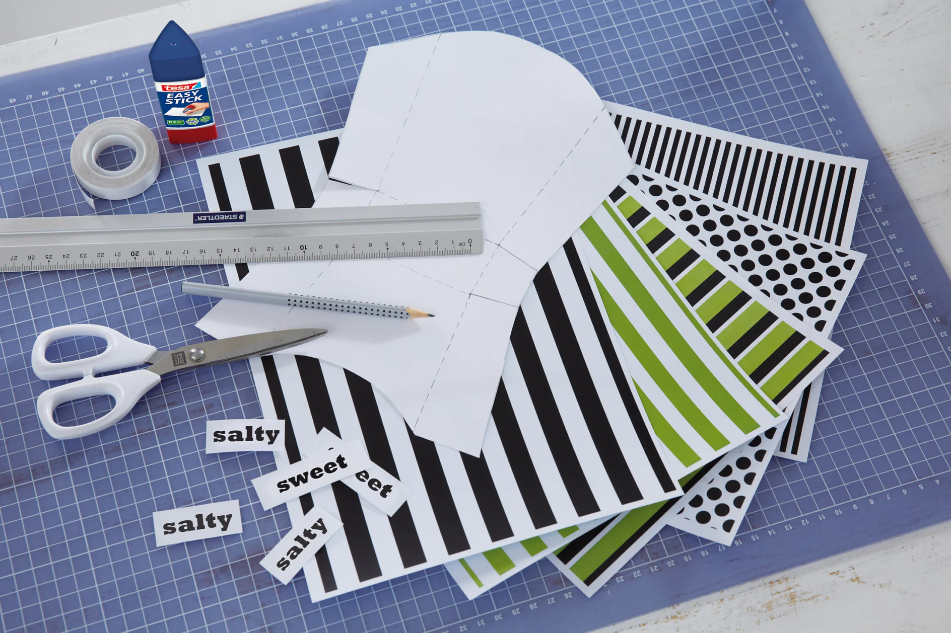 Crafting materials on a table include a pair of scissors, a ruler, a pencil, tesa tape, glue, and sheets of paper with black, white, and green patterns. The papers have polka dots and stripes. The word salty is printed multiple times nearby. (This text has been generated by AI)