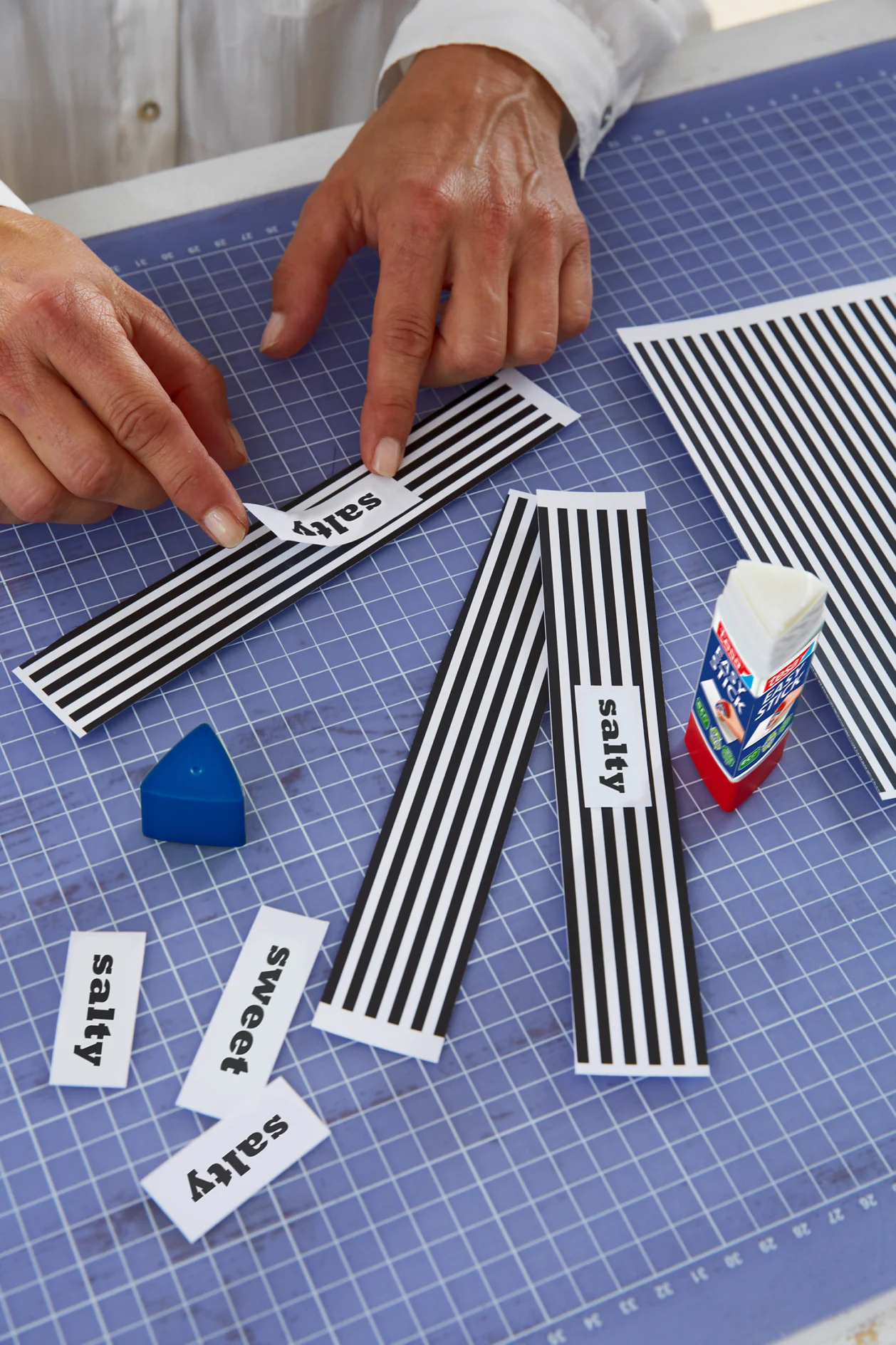 A person wearing a white shirt places a label reading salty onto a striped black and white card. Several similar cards and labels are scattered on a cutting mat, alongside a blue triangular protractor and tesa tape. (This text has been generated by AI)