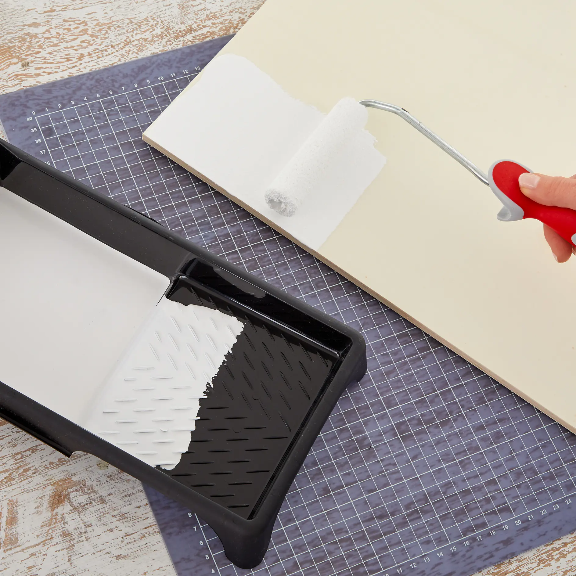 A hand holds a red-handled paint roller applying white paint on a beige surface. Next to it, a black paint tray containing white paint rests on a grid-patterned mat, with tesa tape ensuring precise edges and clean lines throughout the painting process. (This text has been generated by AI)