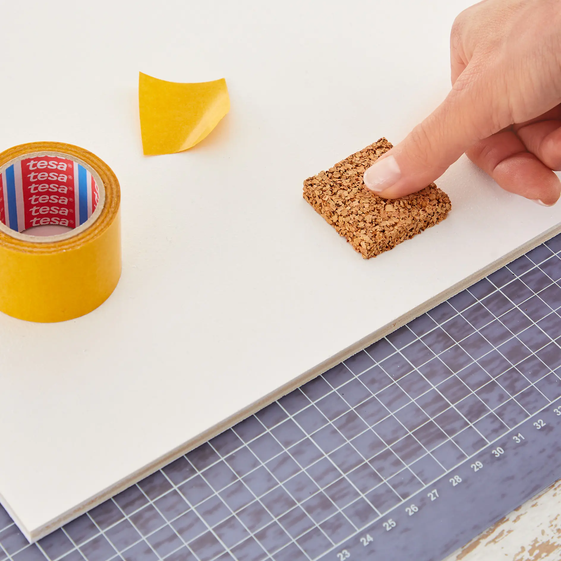 Close-up of a hand holding a small cork square on a white surface, next to a roll of yellow tesa tape and a peeled piece of the same tesa tape. A cutting mat with a grid pattern is partially visible underneath. (This text has been generated by AI)