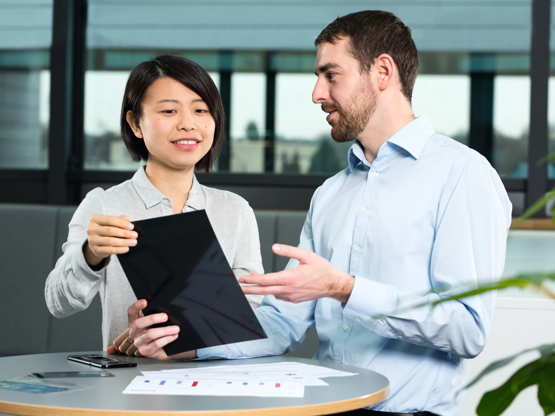 A man and a woman are sitting at a round table, engaged in discussion. The woman holds a tablet, showing it to the man, who gestures with his hand. Papers and tesa tape are on the table. They are in a modern office setting with large windows. (This text has been generated by AI)