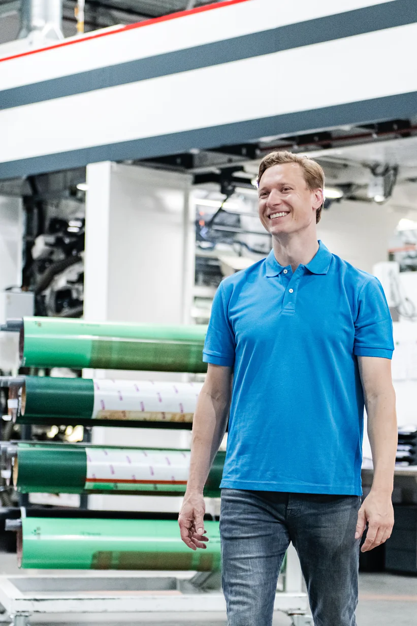 A man in a blue polo shirt and jeans is standing in a large industrial facility. Behind him, there is machinery and large rolls of tesa tape material. The setting appears to be a printing or manufacturing plant. (This text has been generated by AI)