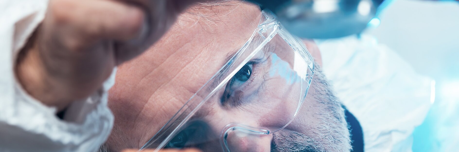A person with safety glasses and a beard is inspecting an electronic circuit board using a magnifying tool. The individual is in close proximity to the components, focusing intently on their work. (This text has been generated by AI)