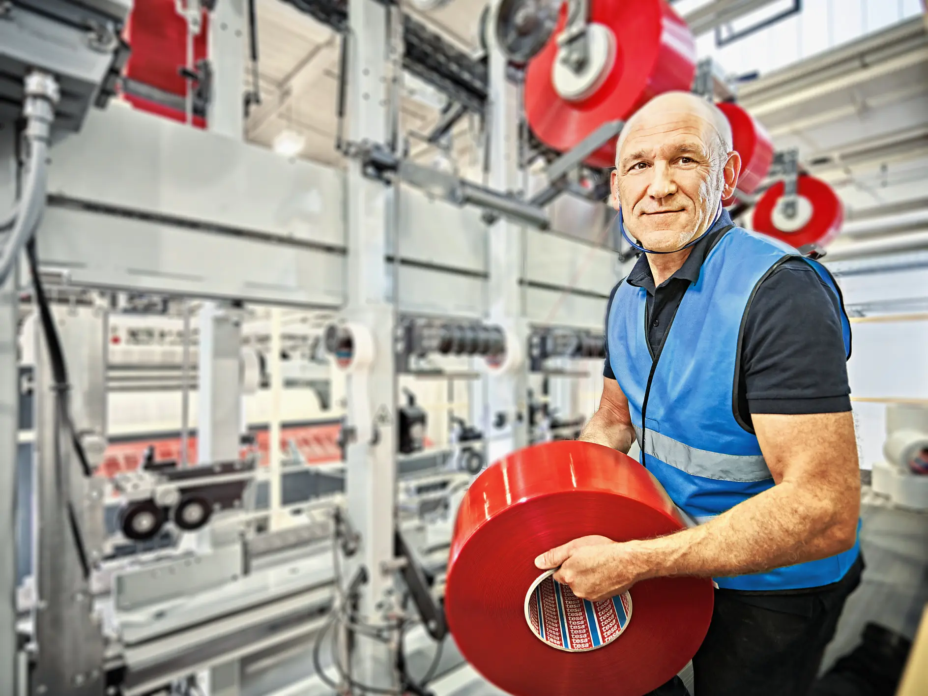 A man in a blue safety vest holds a large red roll of tesa tape in a factory setting. Behind him are industrial machines with more red rolls of tesa tape. The environment is well-lit and industrial.