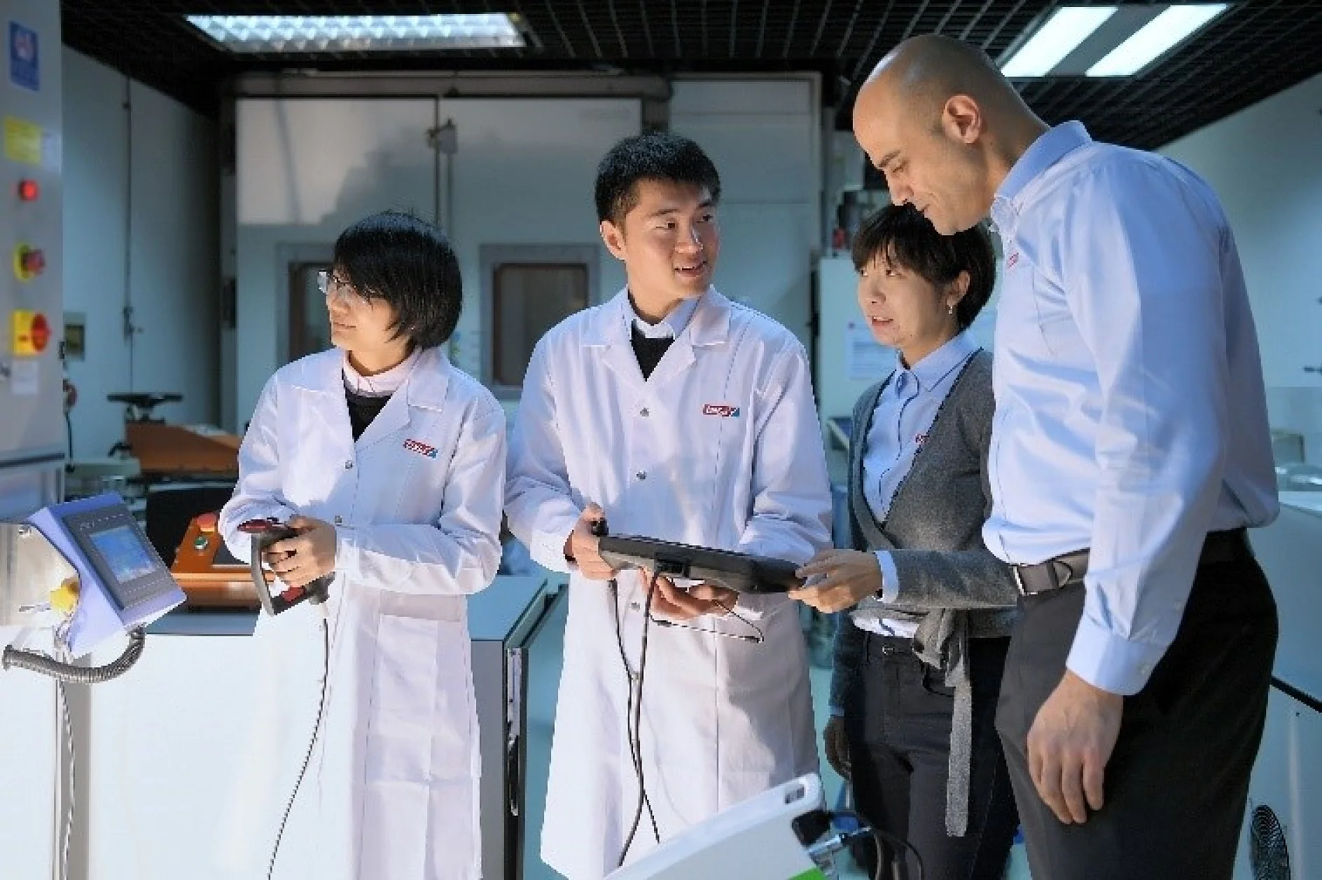 Four people in a laboratory setting, three wearing white lab coats and one in business attire. They are examining electronic devices. The room has various equipment and a digital control panel, along with rolls of tesa tape for securing components during testing and development. (This text has been generated by AI)