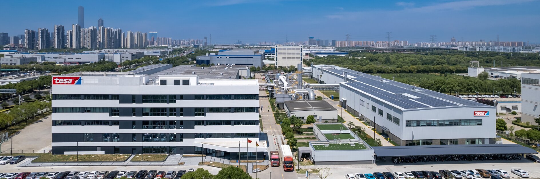 Aerial view of an industrial park with multiple modern buildings under a clear blue sky. The foreground features a parking lot and greenery, while a city skyline is visible in the background. tesa tape logos are displayed on two buildings. (This text has been generated by AI)
