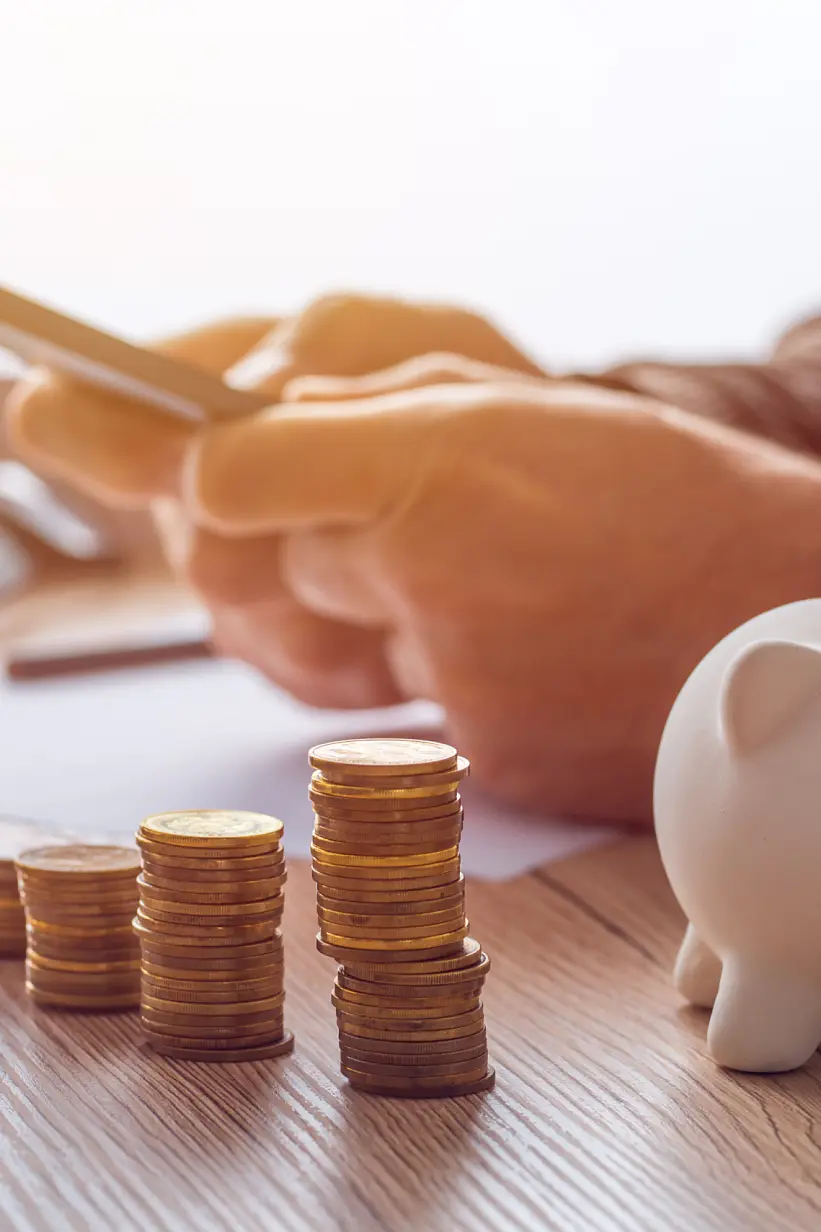 A person uses a smartphone at a desk with neatly stacked coins and a white piggy bank. The scene suggests financial planning or online banking, with a blurred keyboard and papers visible in the background. (This text has been generated by AI)