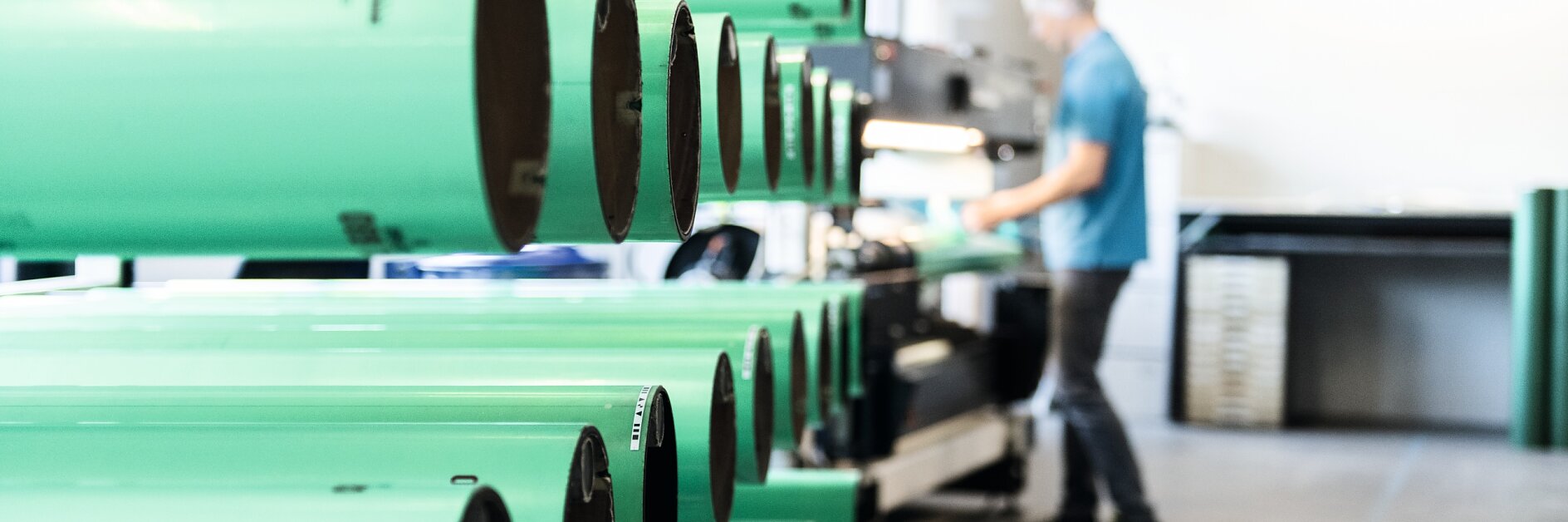 Rows of large green cylindrical tubes are lined up in a workshop. A person wearing a blue shirt is blurred in the background, working with tesa tape and other equipment. The setting appears to be an industrial or manufacturing environment. (This text has been generated by AI)