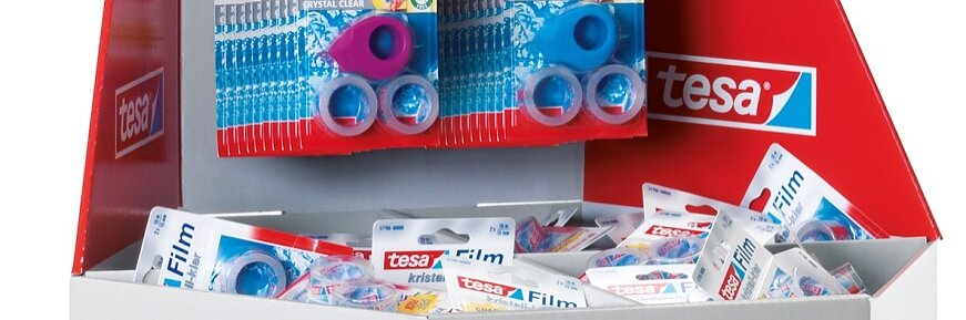 A display stand showcasing various tesa brand school and office supplies, including tesa tape dispensers, correction rollers, and glue sticks. The stand is topped with a small chalkboard illustrating abc and a sun drawing, along with a paper backpack and ruler. (This text has been generated by AI)